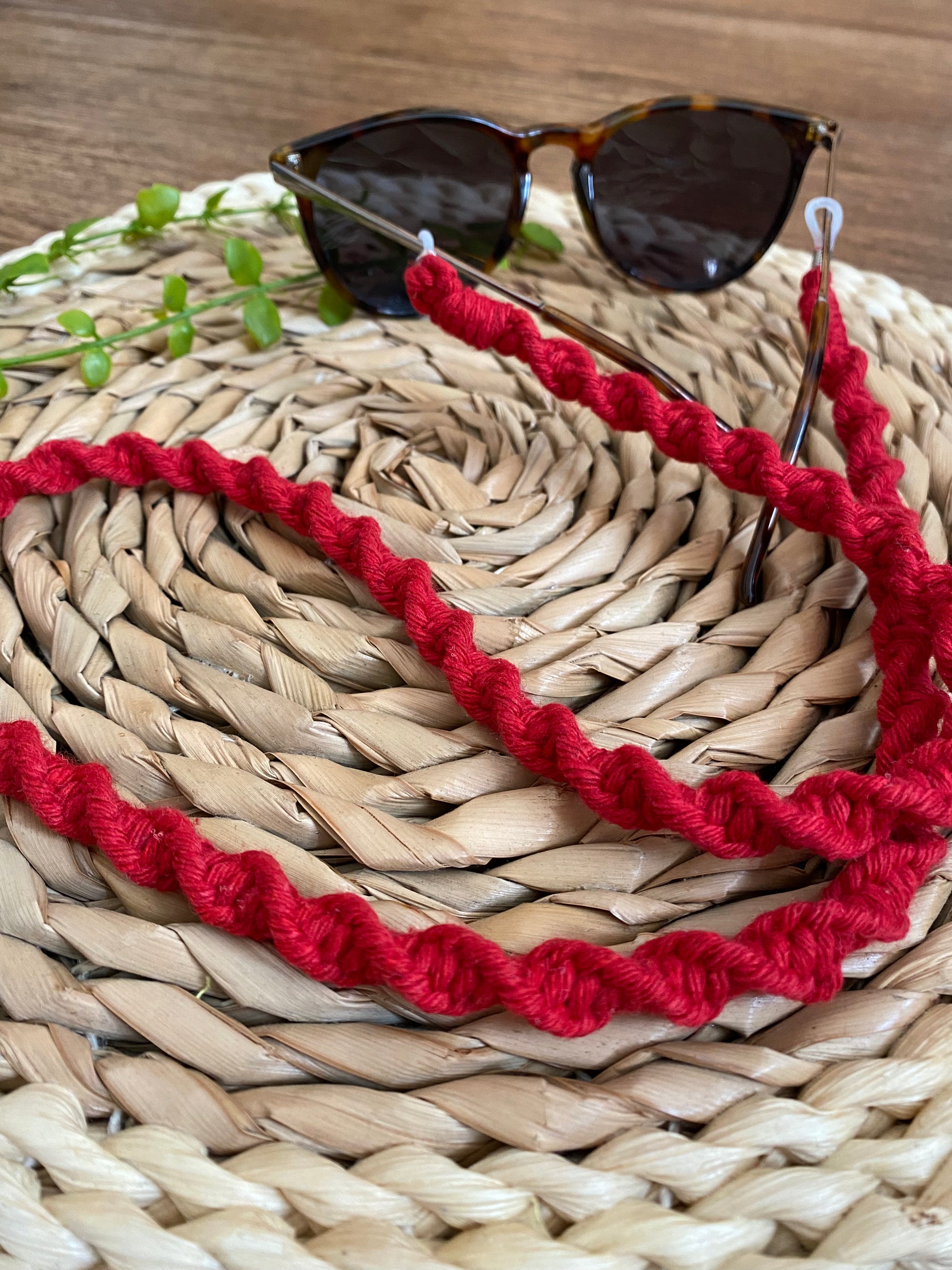 A pair of dark sunglasses are placed on a wicker surface with a handmade macrame eyewear holder chain, made with red recycled cotton yarn, attached to them.