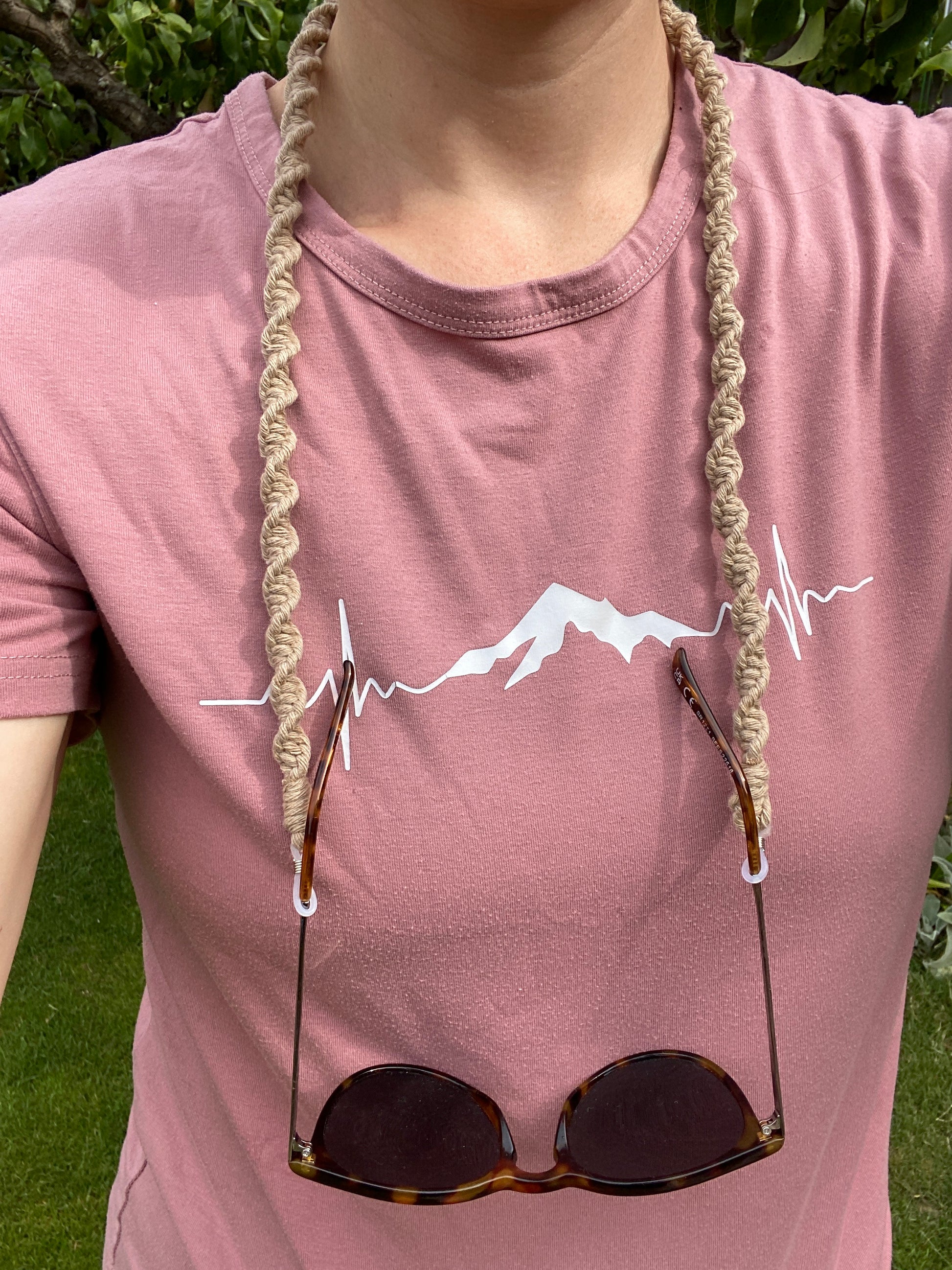 A woman stands outside wearing a pink t shirt with a pair of dark sunglasses hanging from a handmade macrame eyewear holder chain, around her neck.