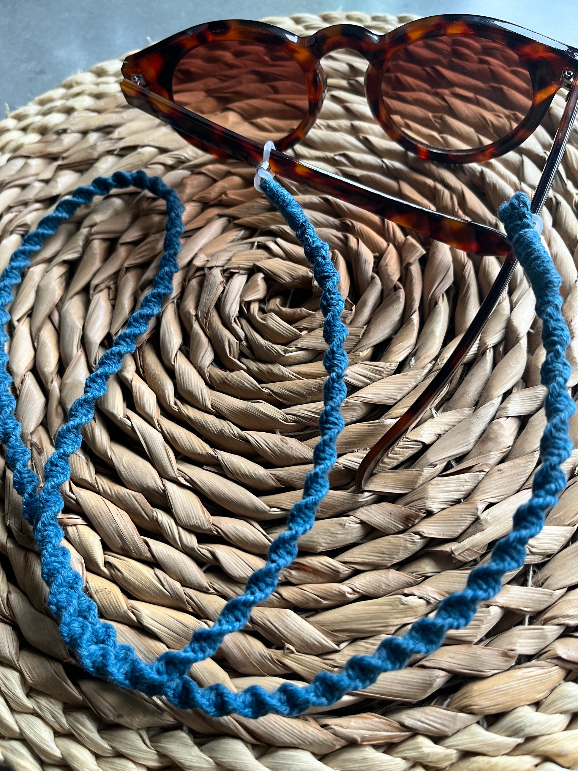 Close-up of tortoiseshell-patterned sunglasses resting on a circular woven placemat. The sunglasses are attached to a teal blue recycled cotton cord spectacles chain.