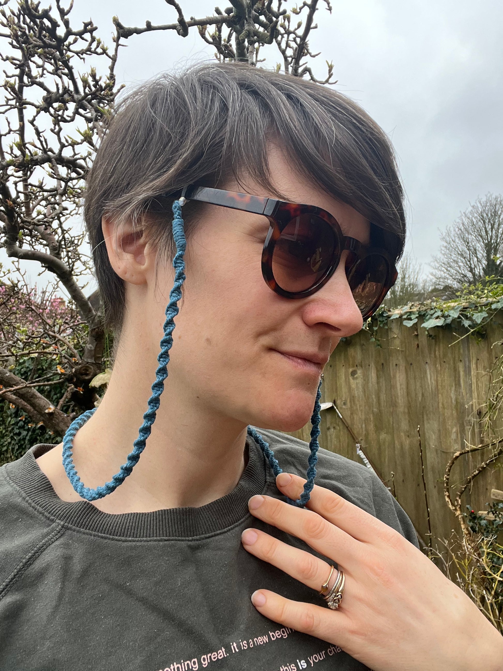 A person with short hair stands outside wearing brown sunglasses, connected to a handmade teal blue recycled cotton eyewear holder chain.