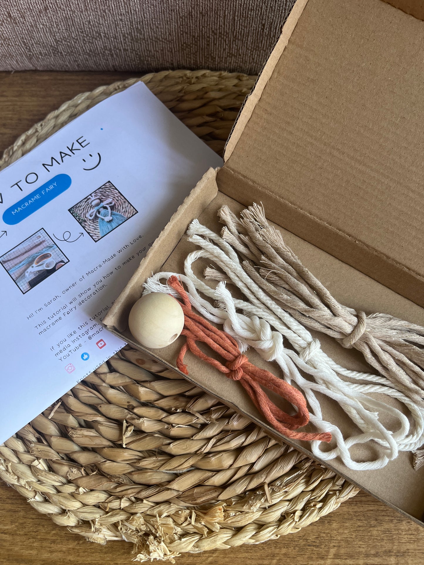 Open cardboard box with macrame yarns and wooden bead inside, with printed DIY craft kit instructions laid beside the box on a wicker surface.