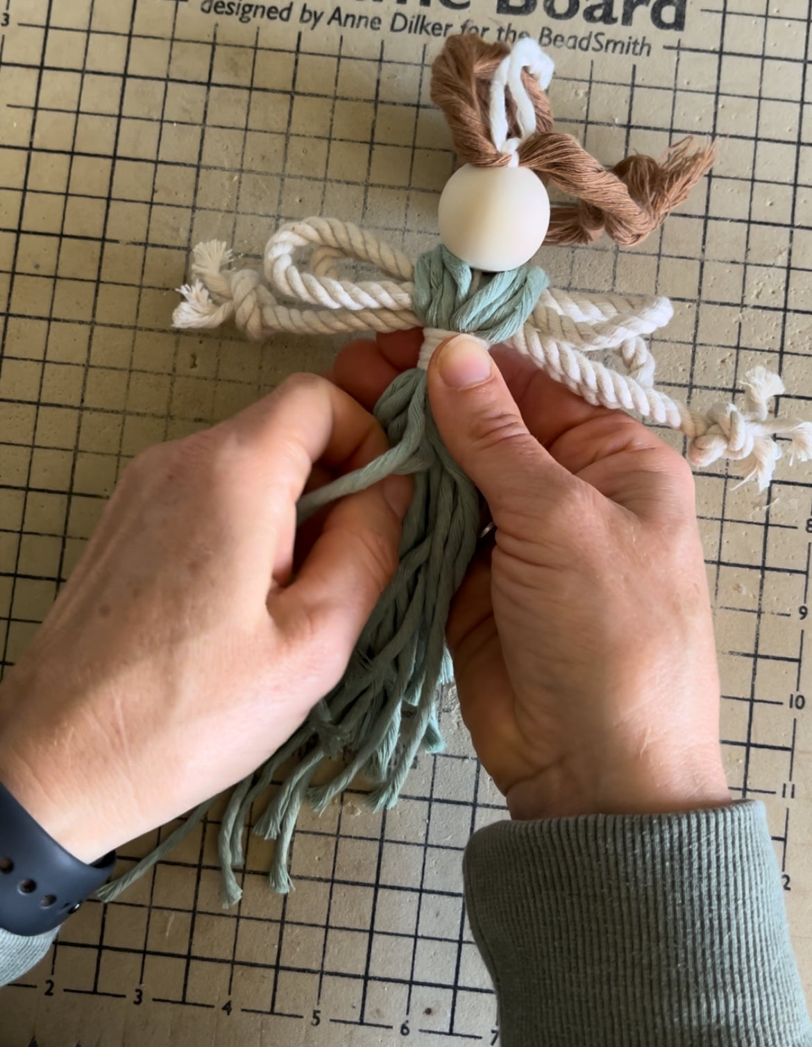 Hands working on a craft project to make a macrame fairy, with yarn and a wooden bead on a grid background