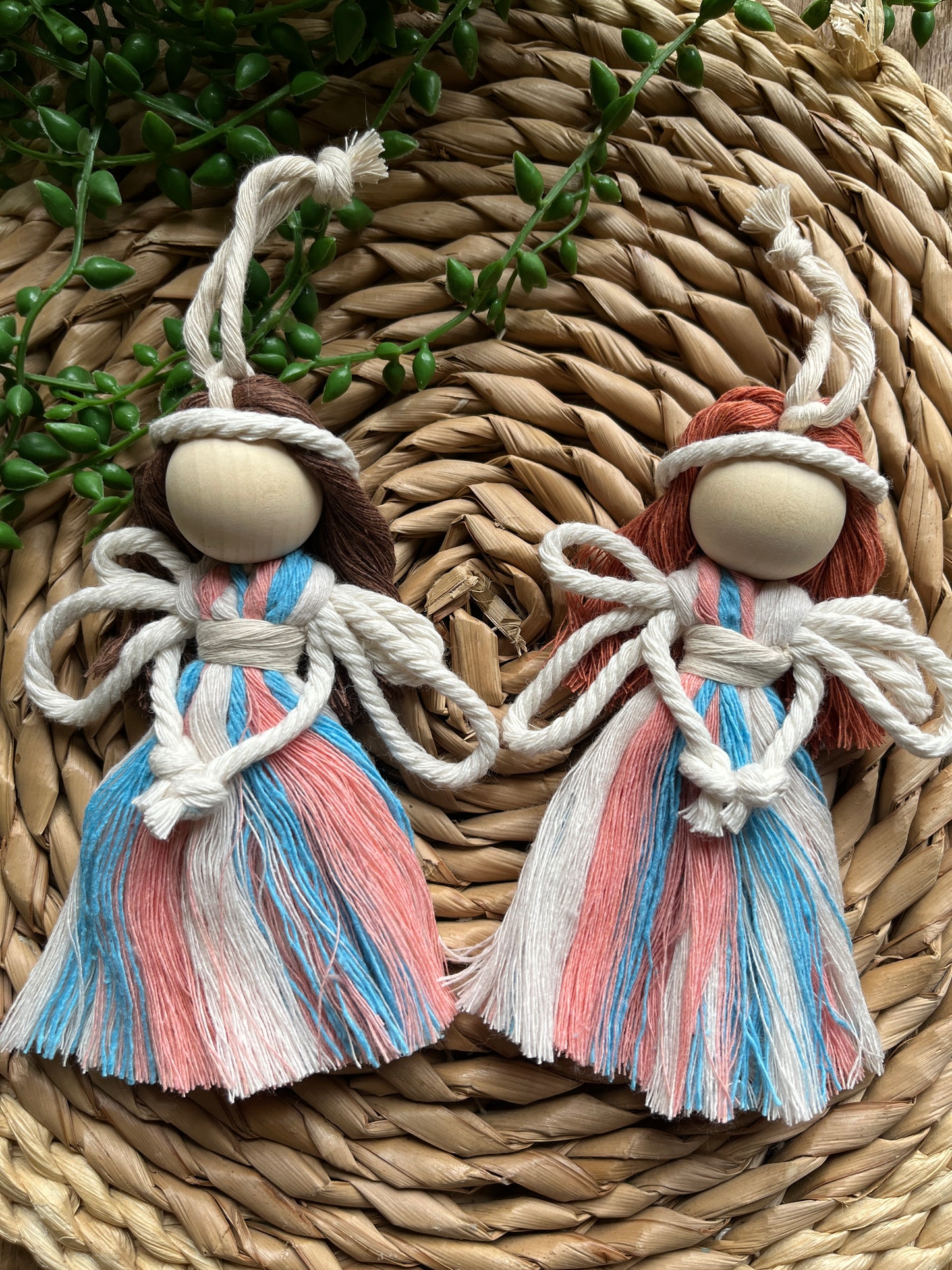 Two handmade macrame fairies, with recycled cotton dresses in blue pink and white, white rope wings and a large wooden bead, laid on a round wicker mat with some green plant vines.
