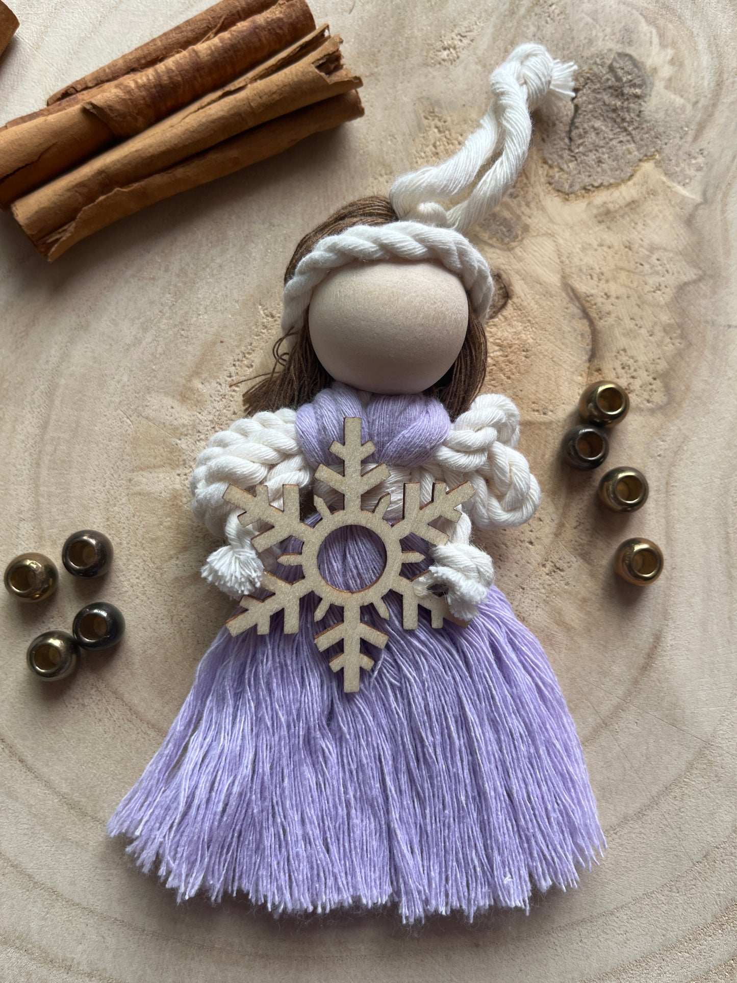 A handmade fairy ornament featuring a lilac dress, white braided rope details, a wooden snowflake, and a wooden bead head, styled on a rustic wood slice background with cinnamon sticks and metal beads.