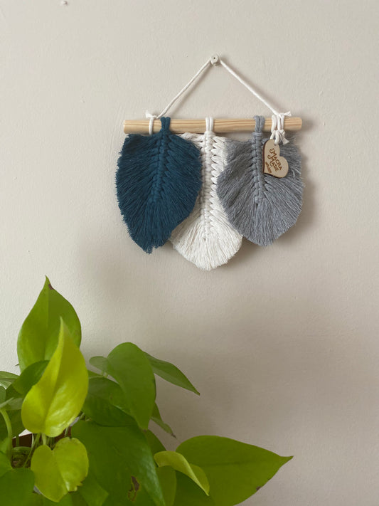 A handmade macrame wall hanging, with three feather shaped accents, made with blue, natural and grey cottons on a wooden dowel rod. A bright green leafy plant is seen in the bottom left corner of the image.
