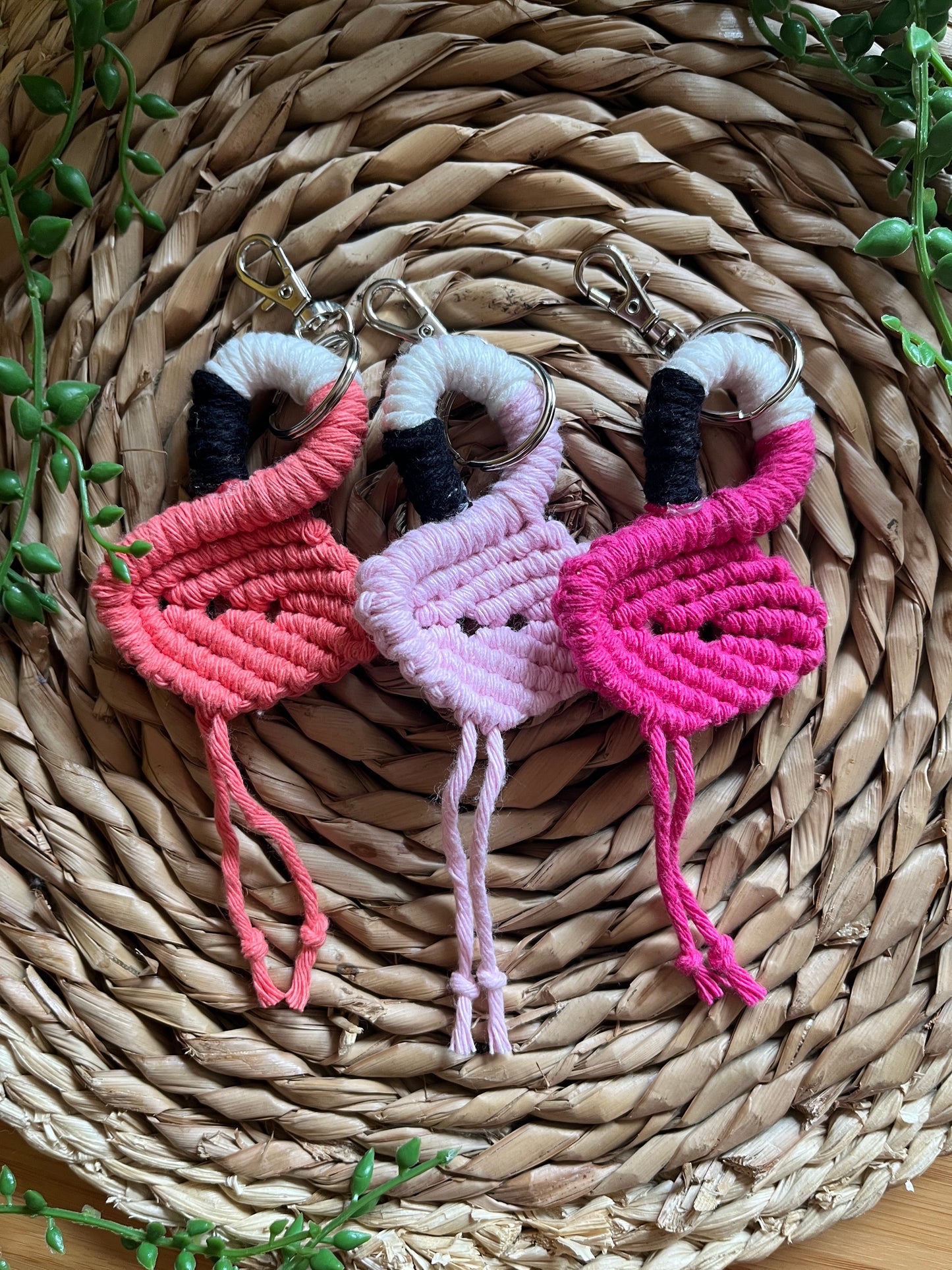 Three handmade macrame keychains in the shape of a flamingos, made using different shades of pink recycled cotton, on a metal split ring with lobster clasp. The keyrings are displayed on a round wicker surface next to some green plant vines.