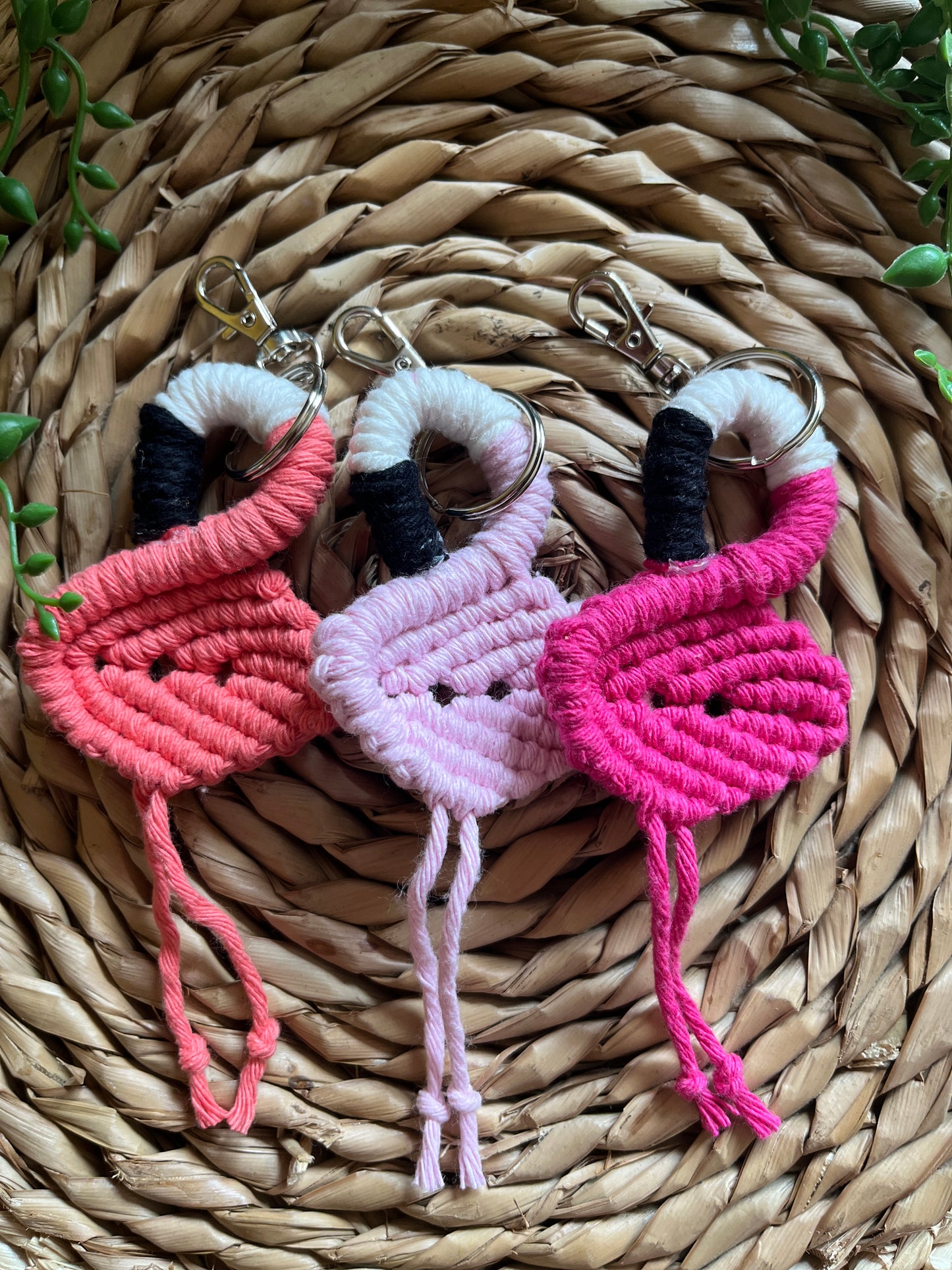 Three handmade macrame keychains in the shape of a flamingos, made using different shades of pink recycled cotton, on a metal split ring with lobster clasp. The keyrings are displayed on a round wicker surface next to some green plant vines.