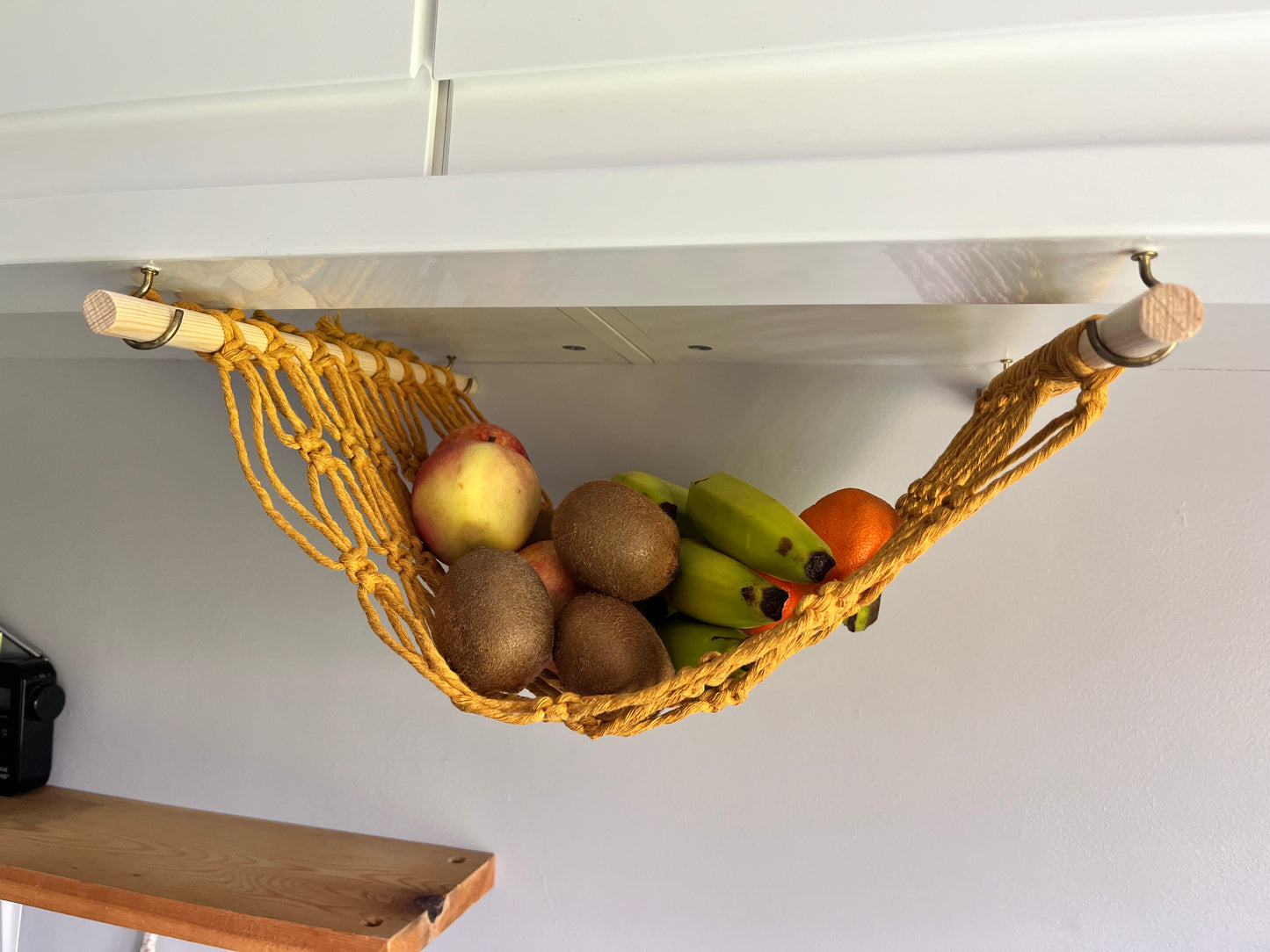 Various fruits hang in a macrame hammock made with mustard yellow recycled cotton and wooden dowels, underneath a white kitchen wall unit.