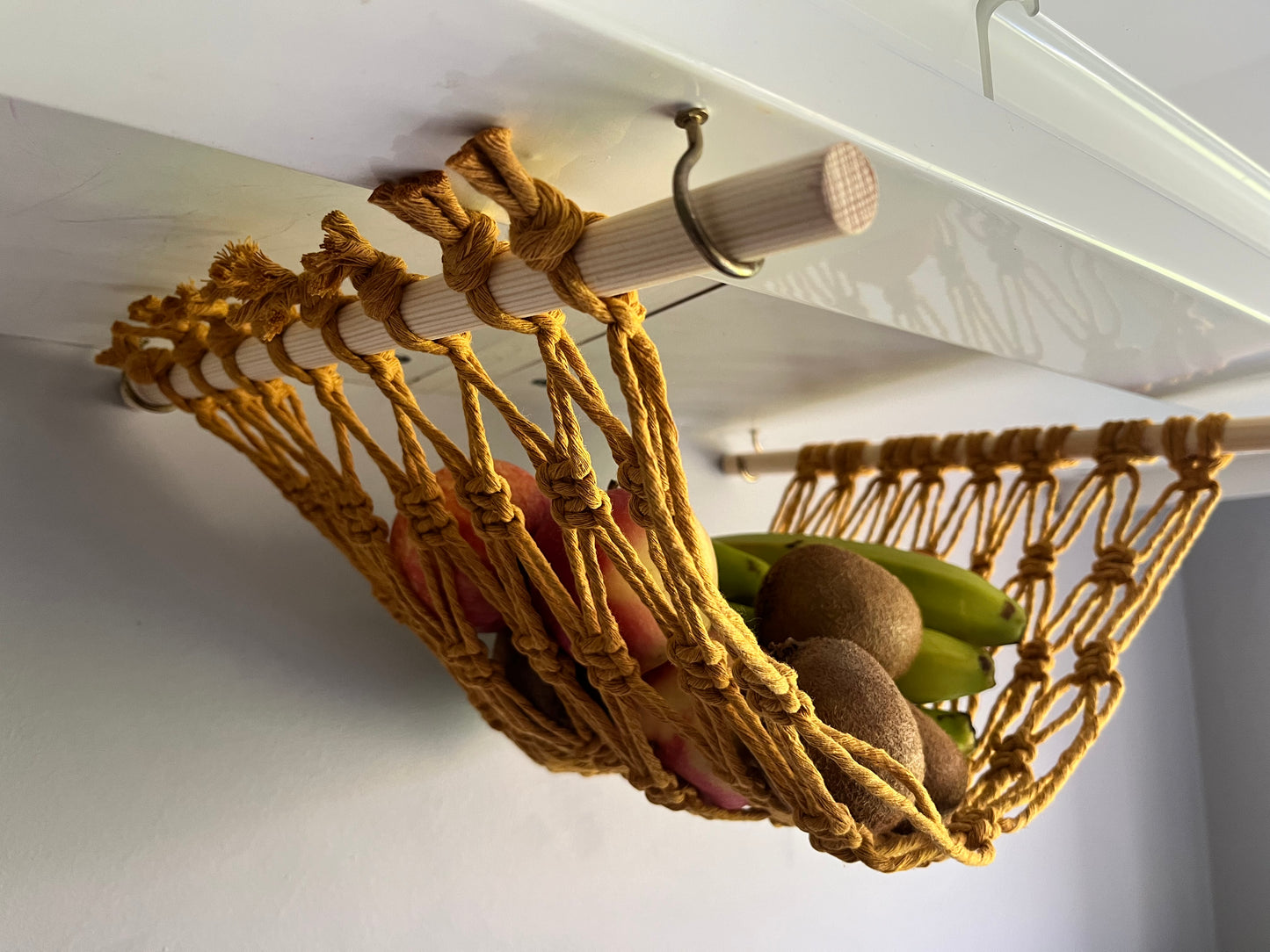 Close up of a macrame hammock made with mustard yellow recycled cotton and wooden dowels, holding fruit underneath a white kitchen wall unit.