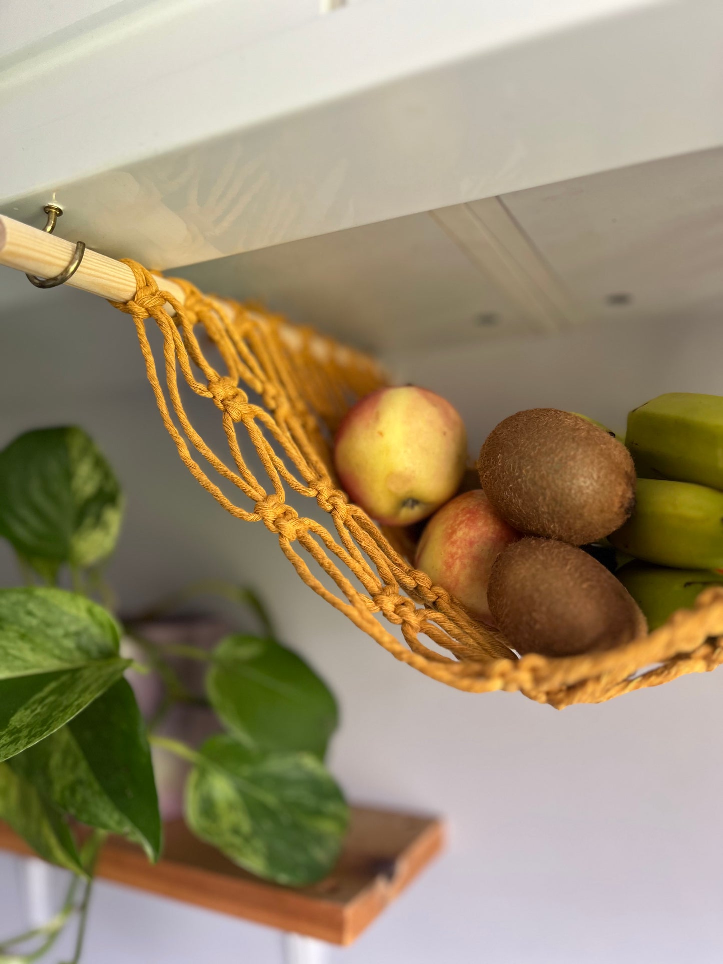 Kiwis and apples hang in a macrame hammock made with mustard yellow recycled cotton and wooden dowels, underneath a white kitchen wall unit. There is a trailing green plant in the background.