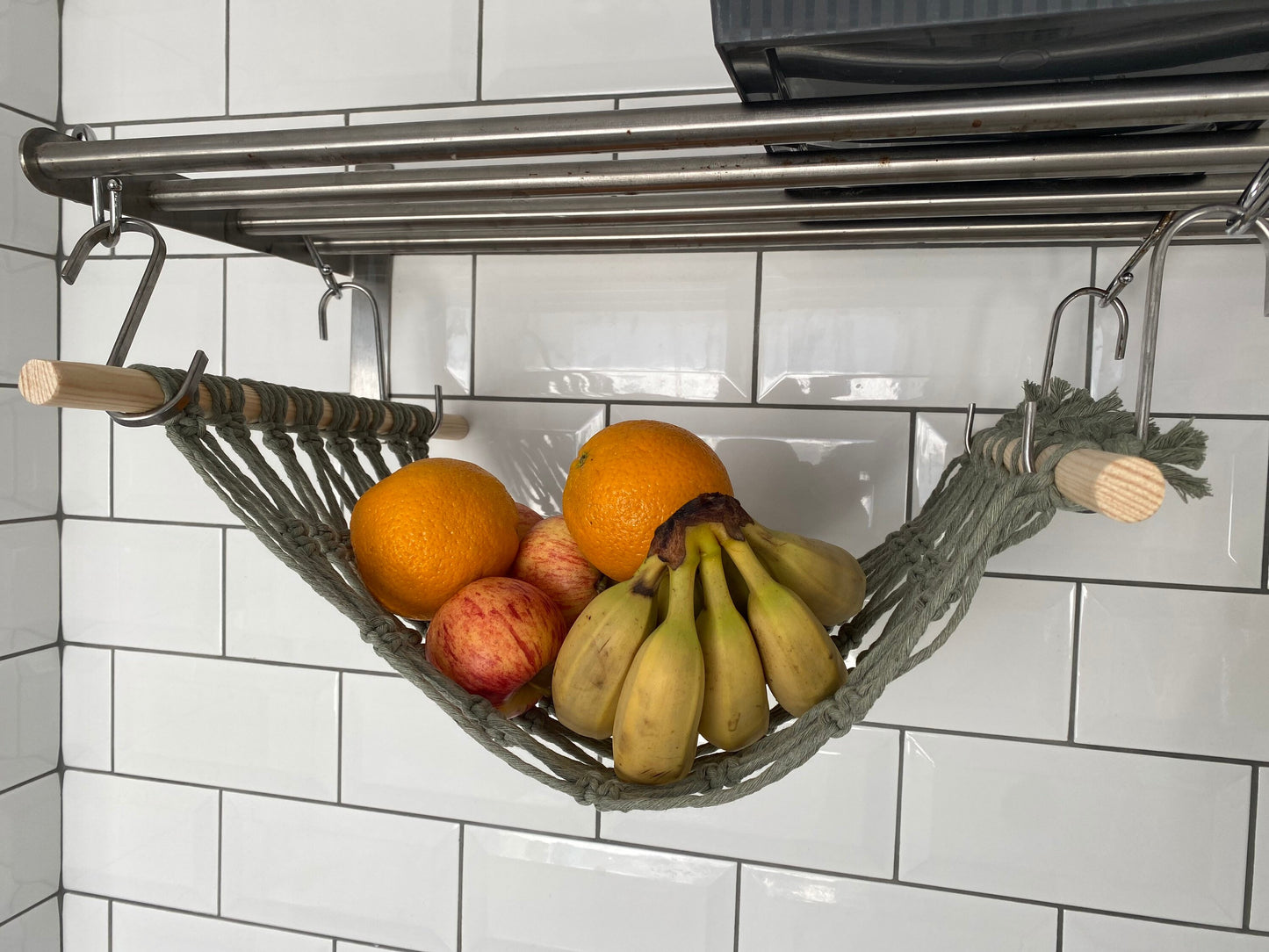 Bananas, oranges and apples hang in a macrame hammock made with sage green recycled cotton and wooden dowels, underneath a metal wall shelf from S hooks.