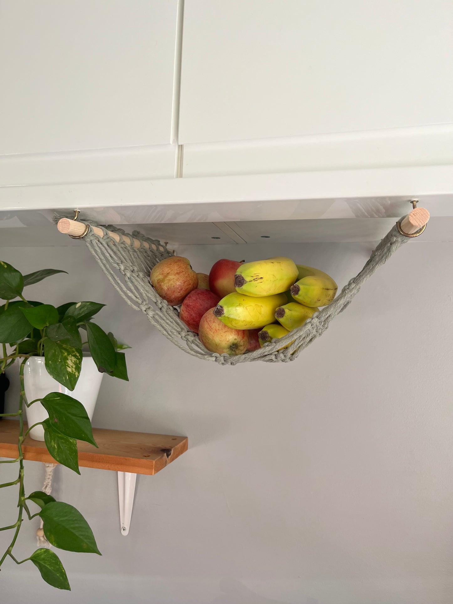 Bananas and apples hang in a macrame hammock made with sage green recycled cotton and wooden dowels, underneath a white kitchen wall unit. There is a trailing green plant on a wooden shelf in the background.