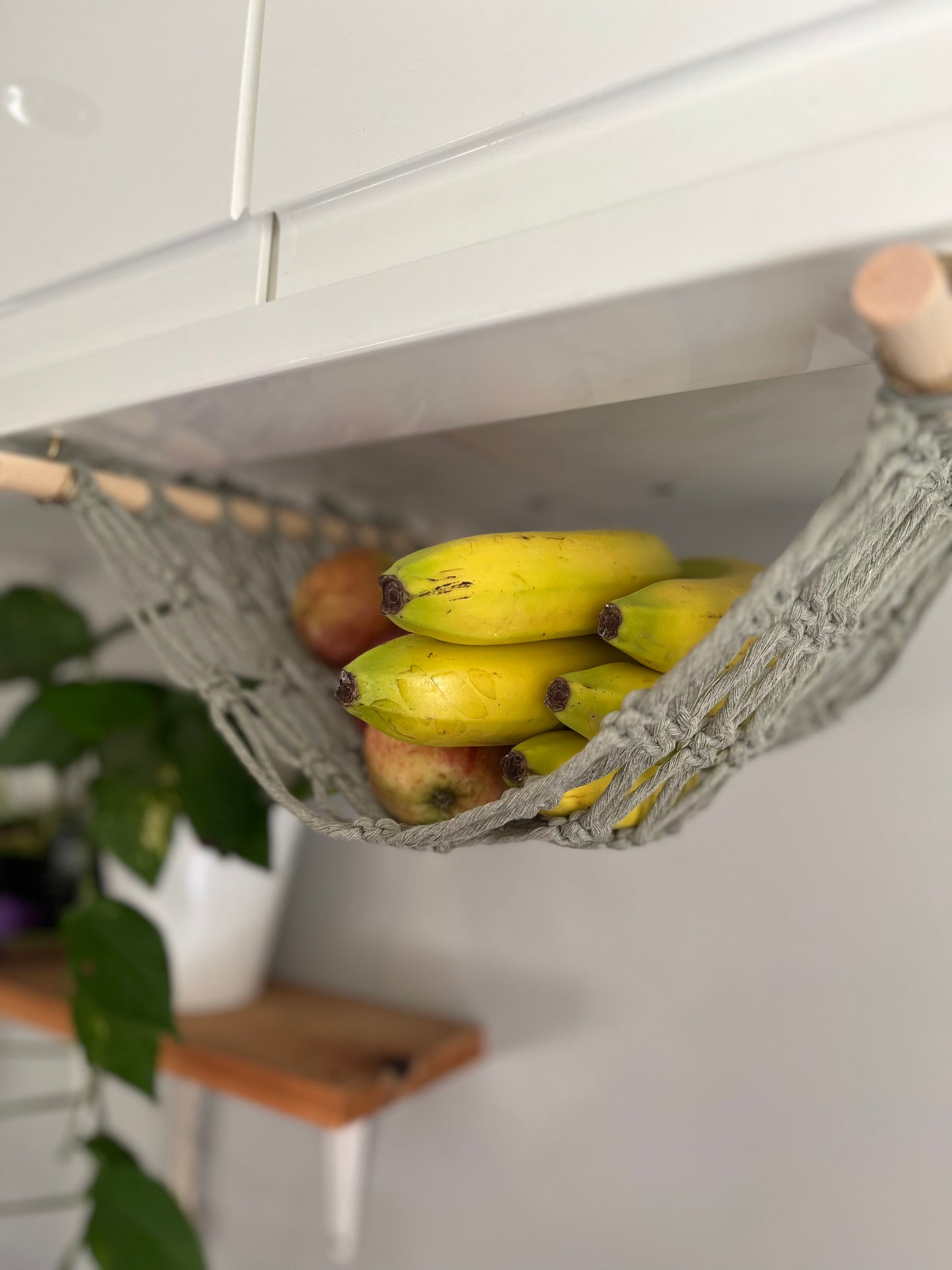 Close up of bananas in a macrame hammock made with sage green recycled cotton and wooden dowels, hanging underneath a white kitchen wall unit.
