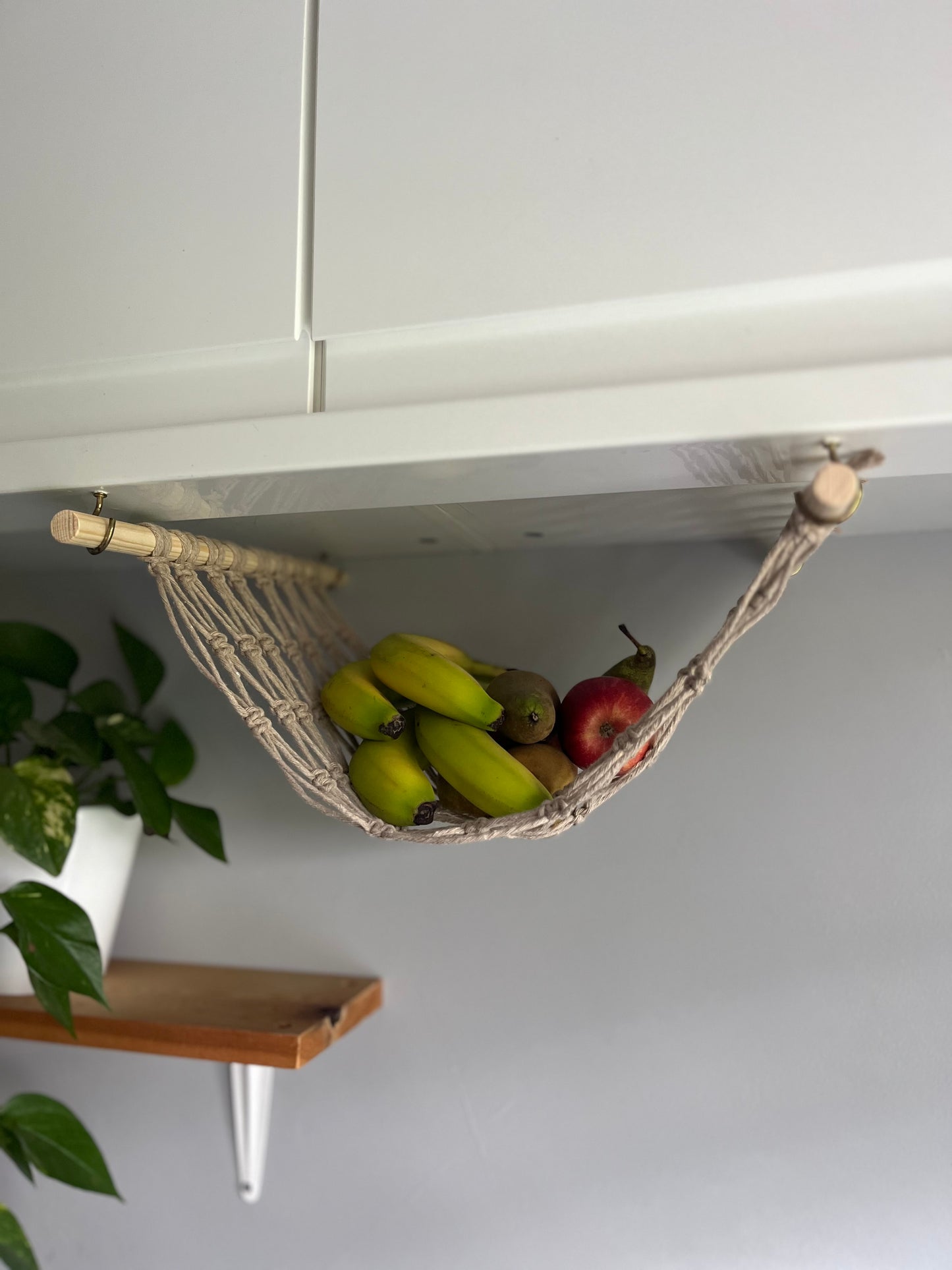 Bananas and apples hang in a macrame hammock made with taupe recycled cotton and wooden dowels, underneath a white kitchen wall unit. There is a trailing green plant on a wooden shelf in the background.