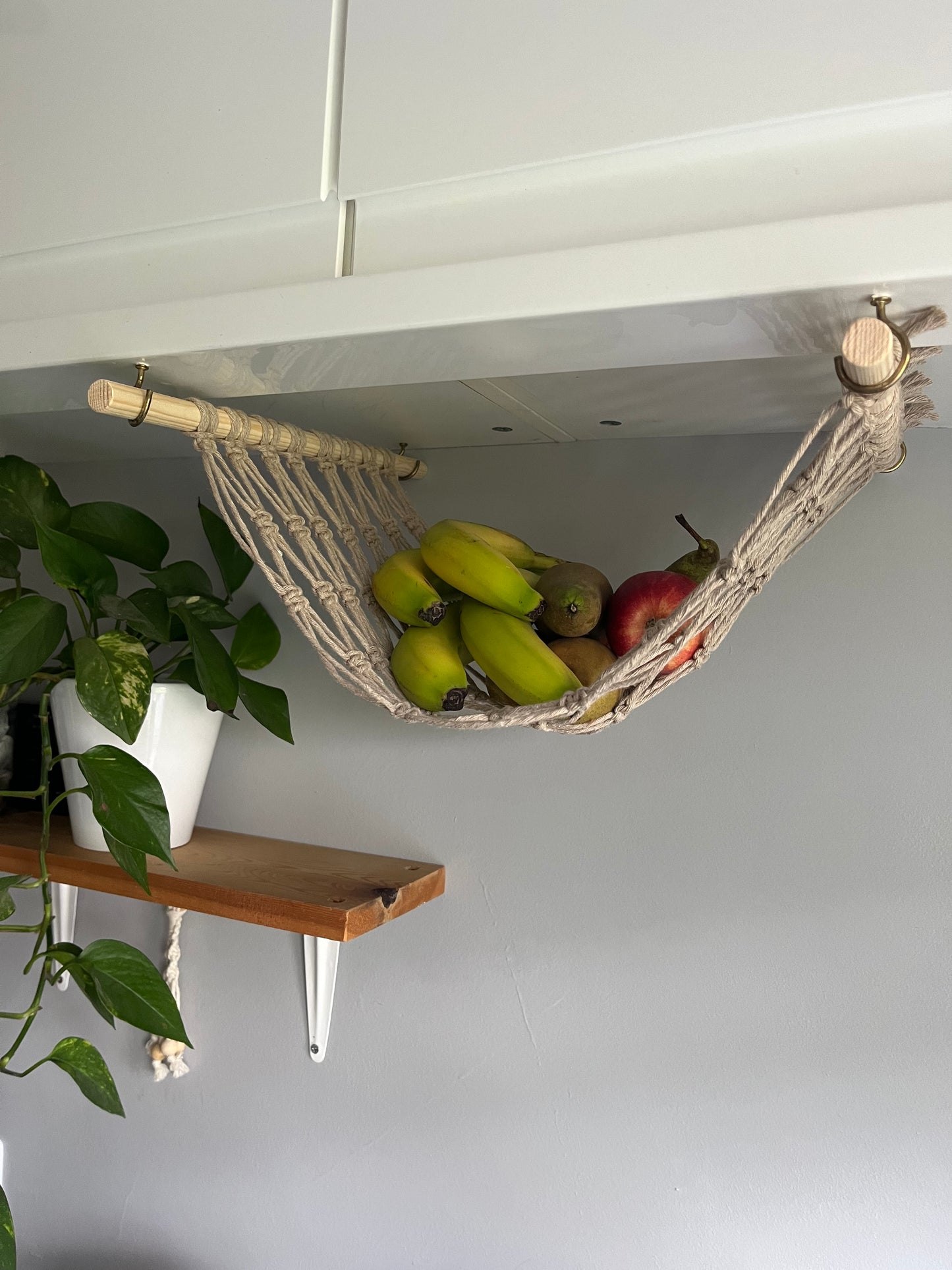 Bananas and apples hang in a macrame hammock made with taupe recycled cotton and wooden dowels, underneath a white kitchen wall unit.