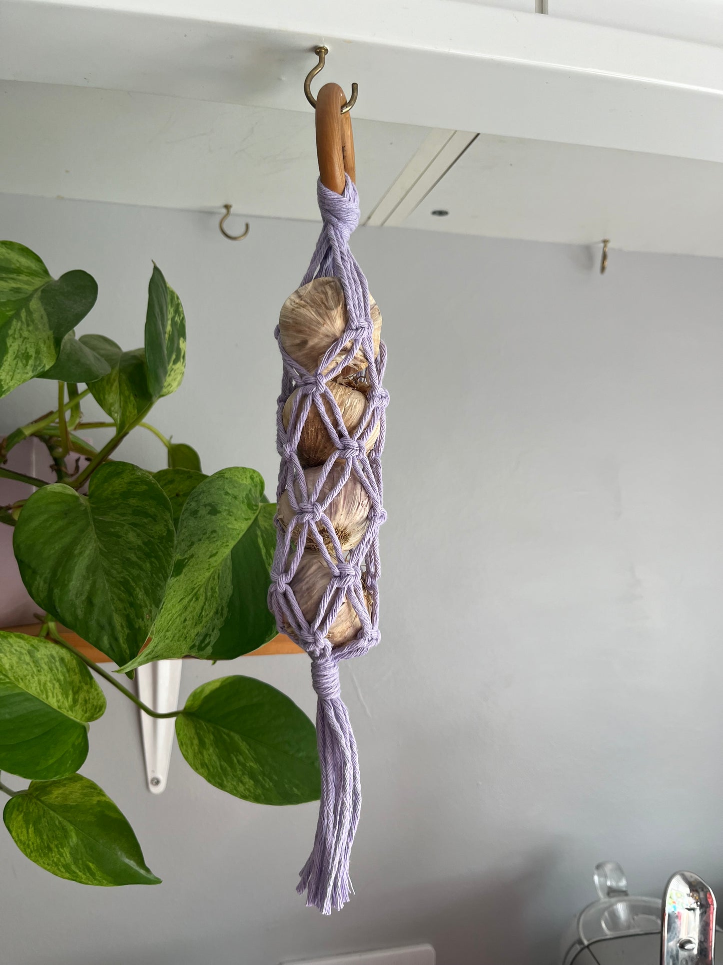 A handmade macrame garlic hanger made with lilac cotton yarn and a wooden ring, holding 4 fresh garlic bulbs, hanging under a kitchen cabinet with a leafy green houseplant in the background.
