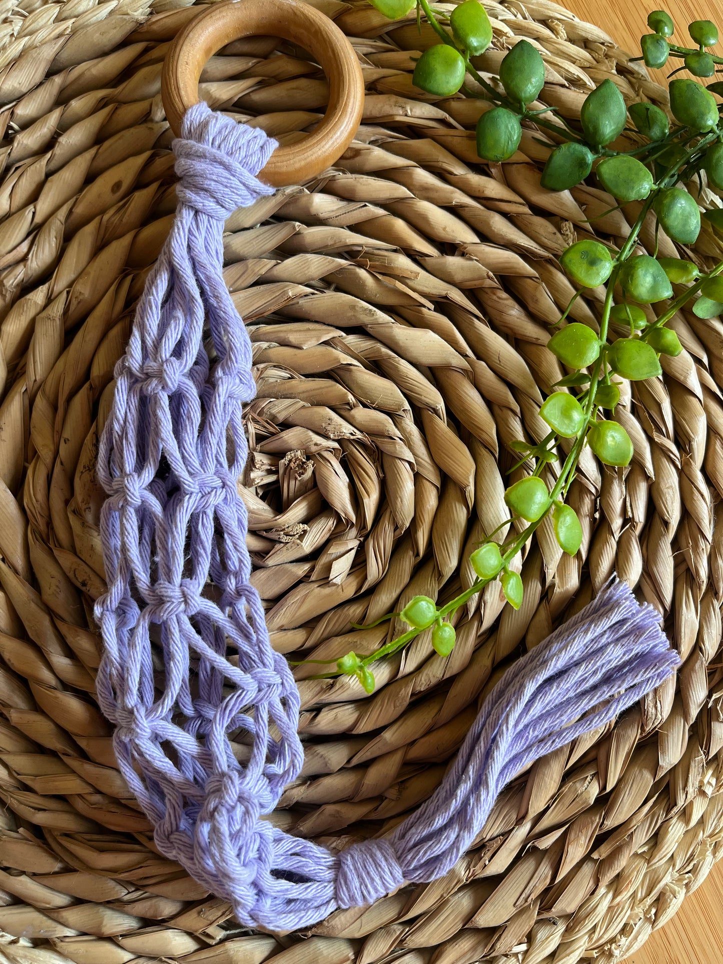 A handmade macrame garlic hanger made with lilac cotton yarn and a wooden ring, folded neatly on a round wicker placemat with some green plant vines.