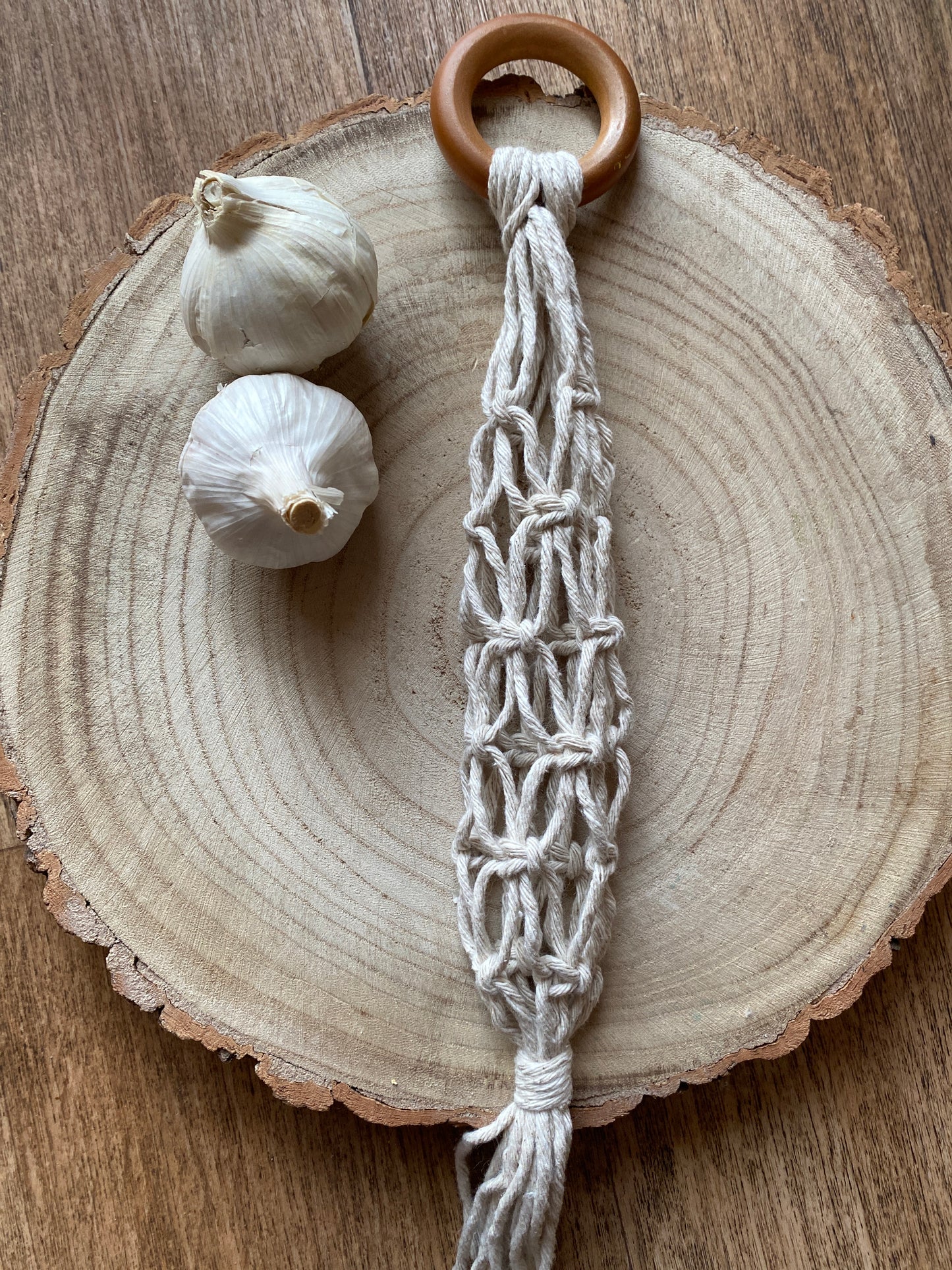 A handmade macrame garlic hanger made with natural cotton yarn and a wooden ring, laid on a round wooden board next to two fresh garlic bulbs.