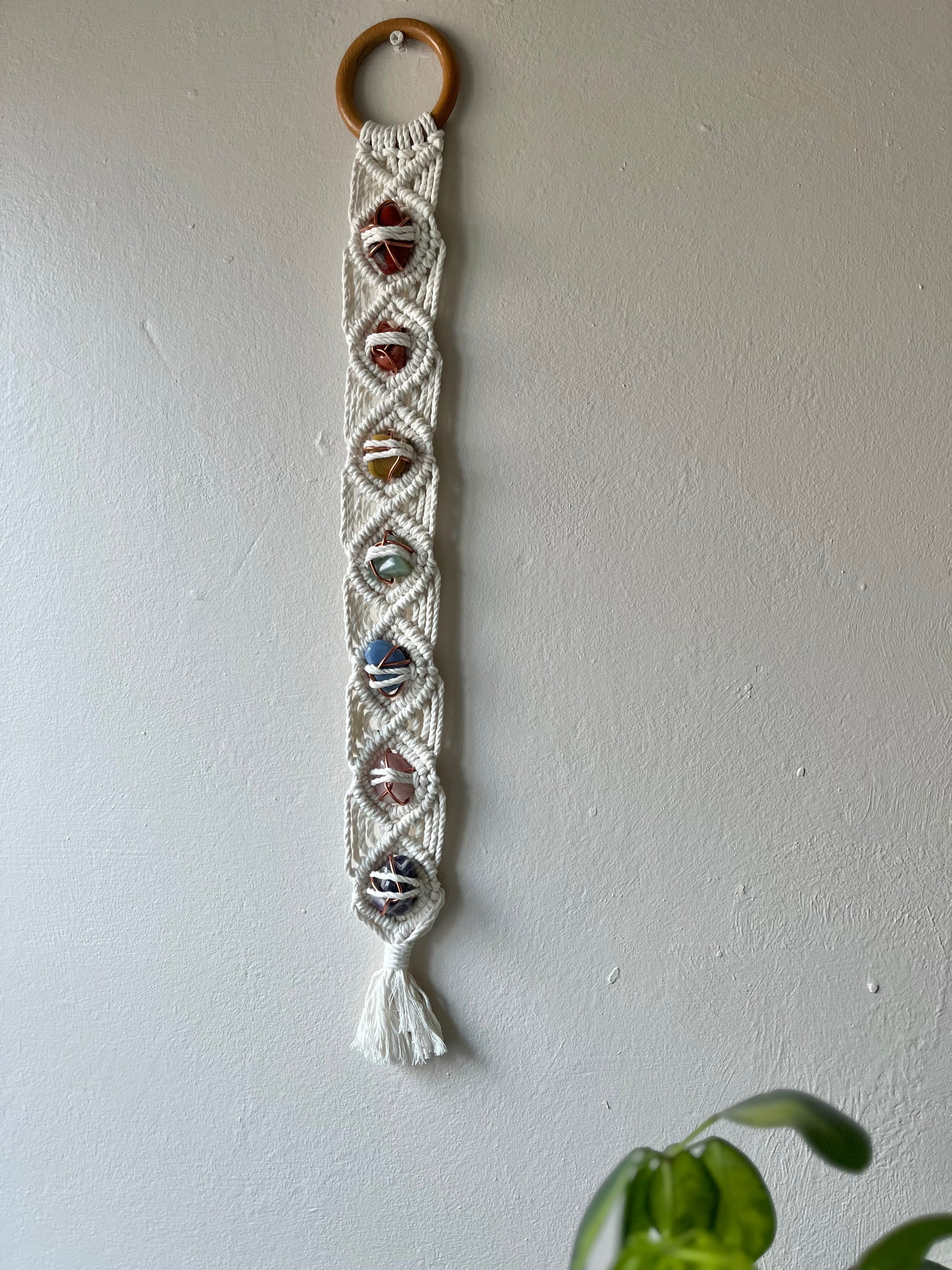 Long macrame wall hanging, featuring 7 gemstones to represent the chakras, held together with copper wire, and wrapped in natural recycled cotton yarn using a diamond knot pattern. finished with a repurposed wooden hanging ring. Displayed hanging on a white painted wall.