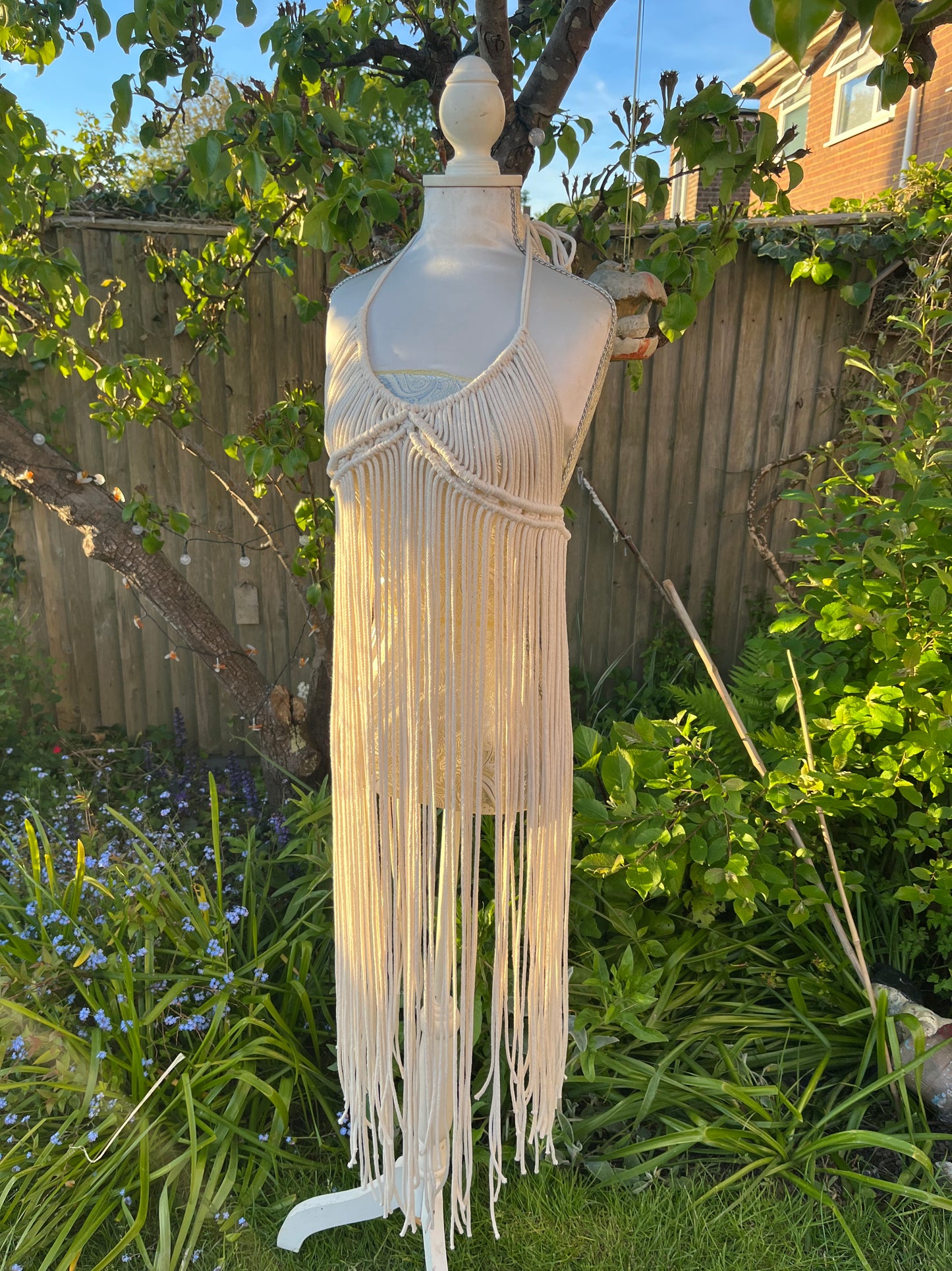A beige, one-size macrame boho goddess dress with long fringe on a mannequin outdoors.