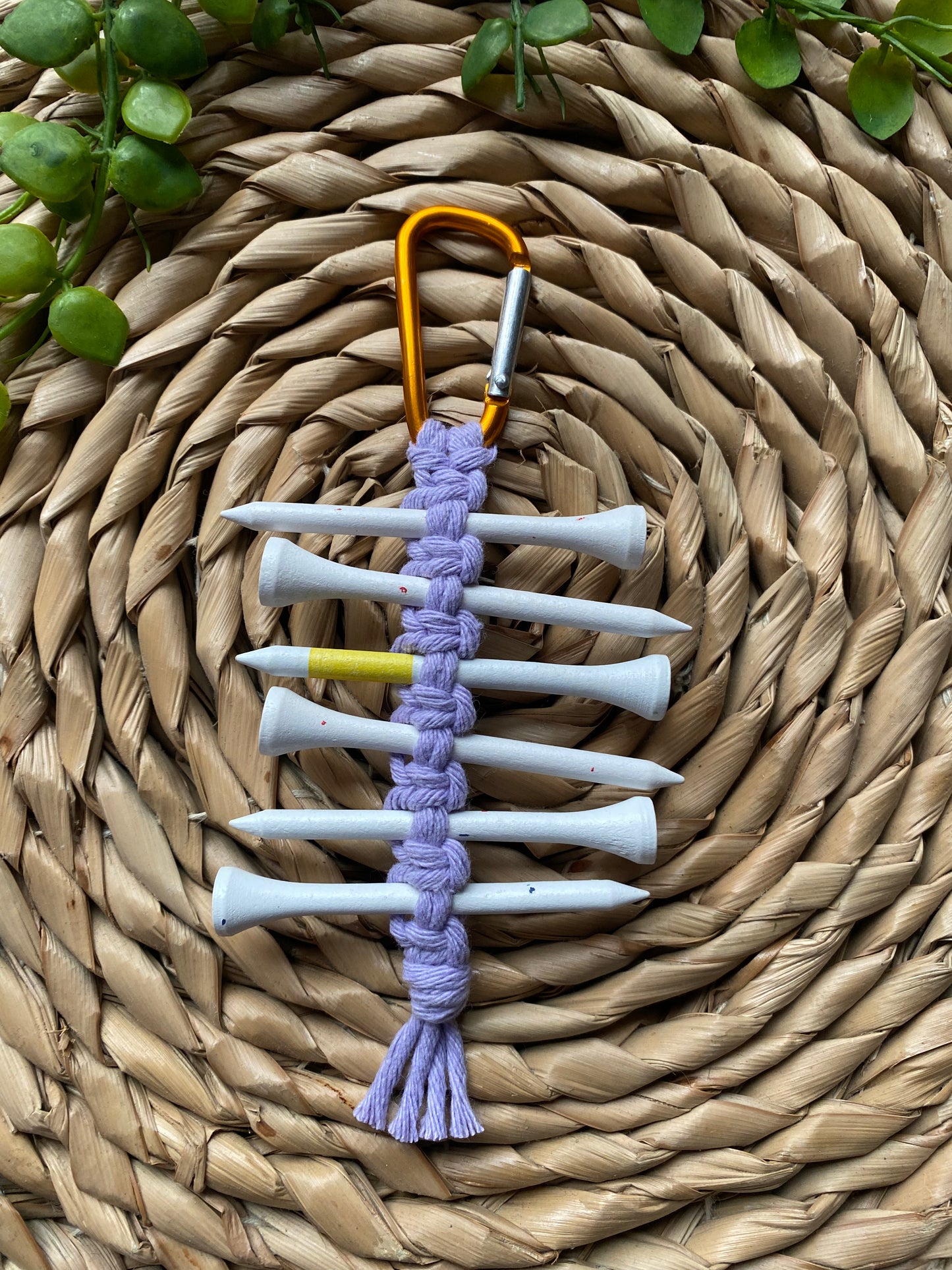 A knotted golf tee holder, made with lilac yarn on an orange gold carabiner clip, laid on a wicker surface holding 6 white golf tees.