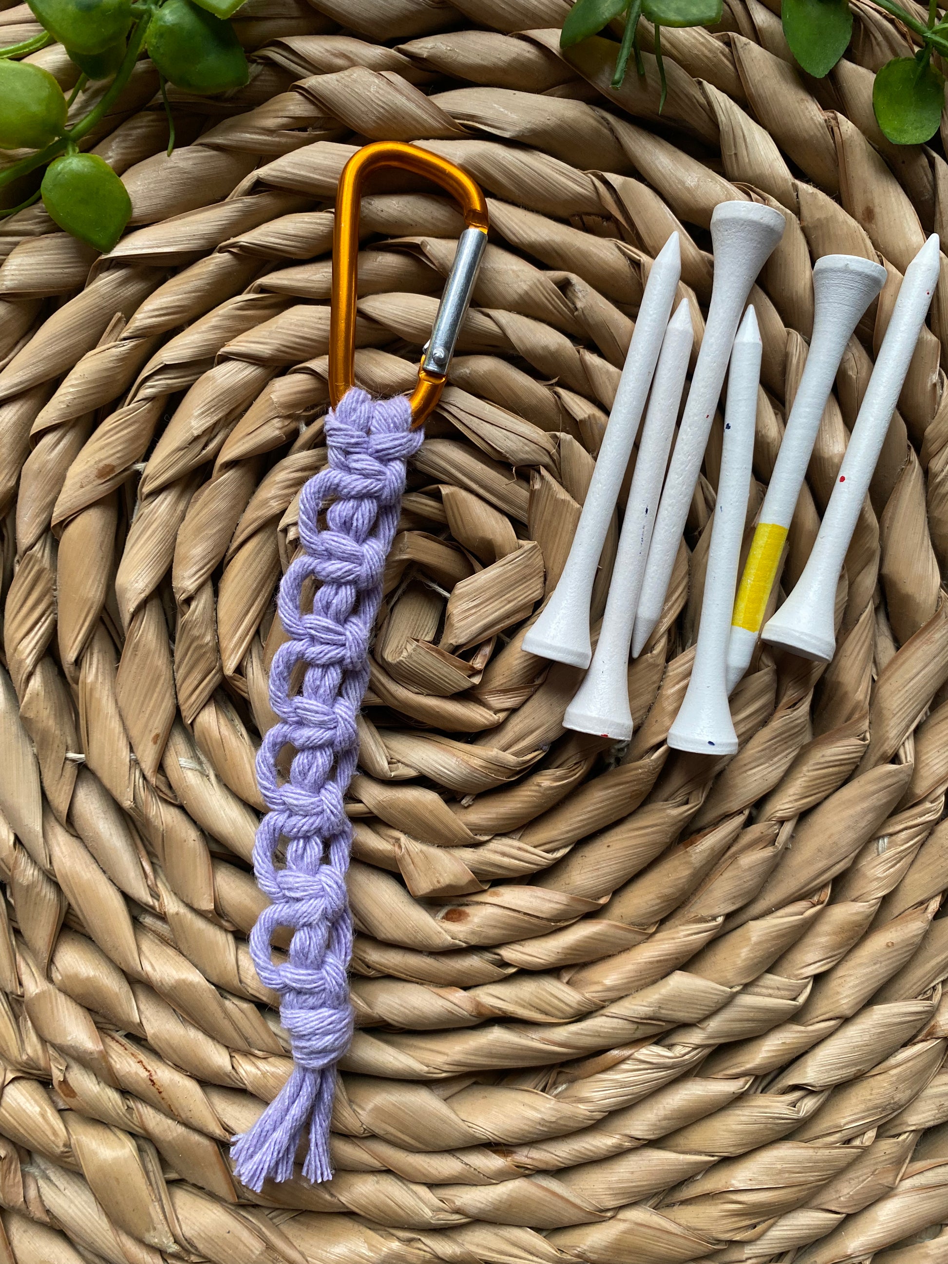A knotted golf tee holder, made with lilac yarn on an orange gold carabiner clip, laid on a wicker surface next to 6 white golf tees.