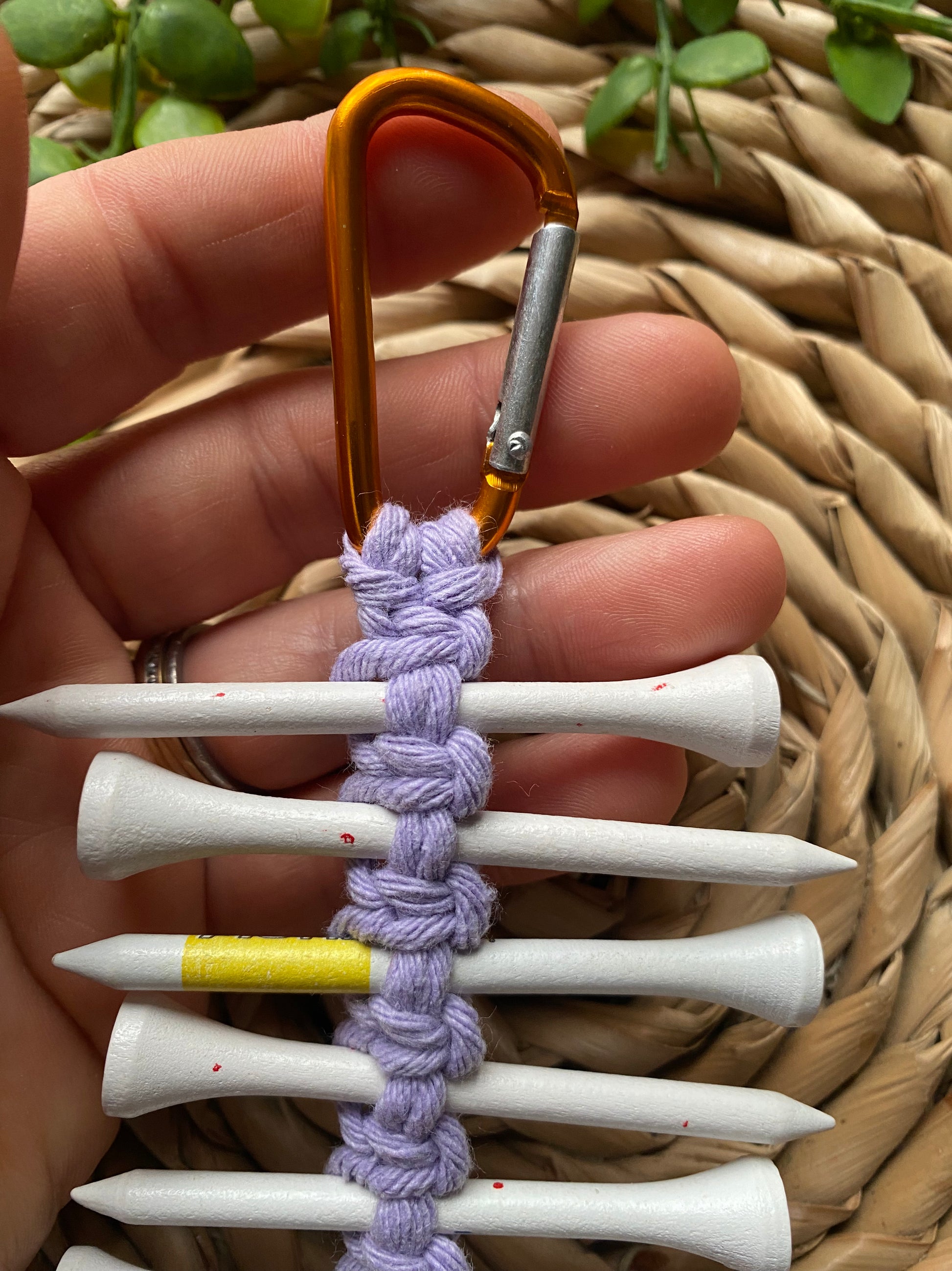A close up of a hand holding a knotted golf tee holder, made with lilac yarn on an orange gold carabiner clip.
