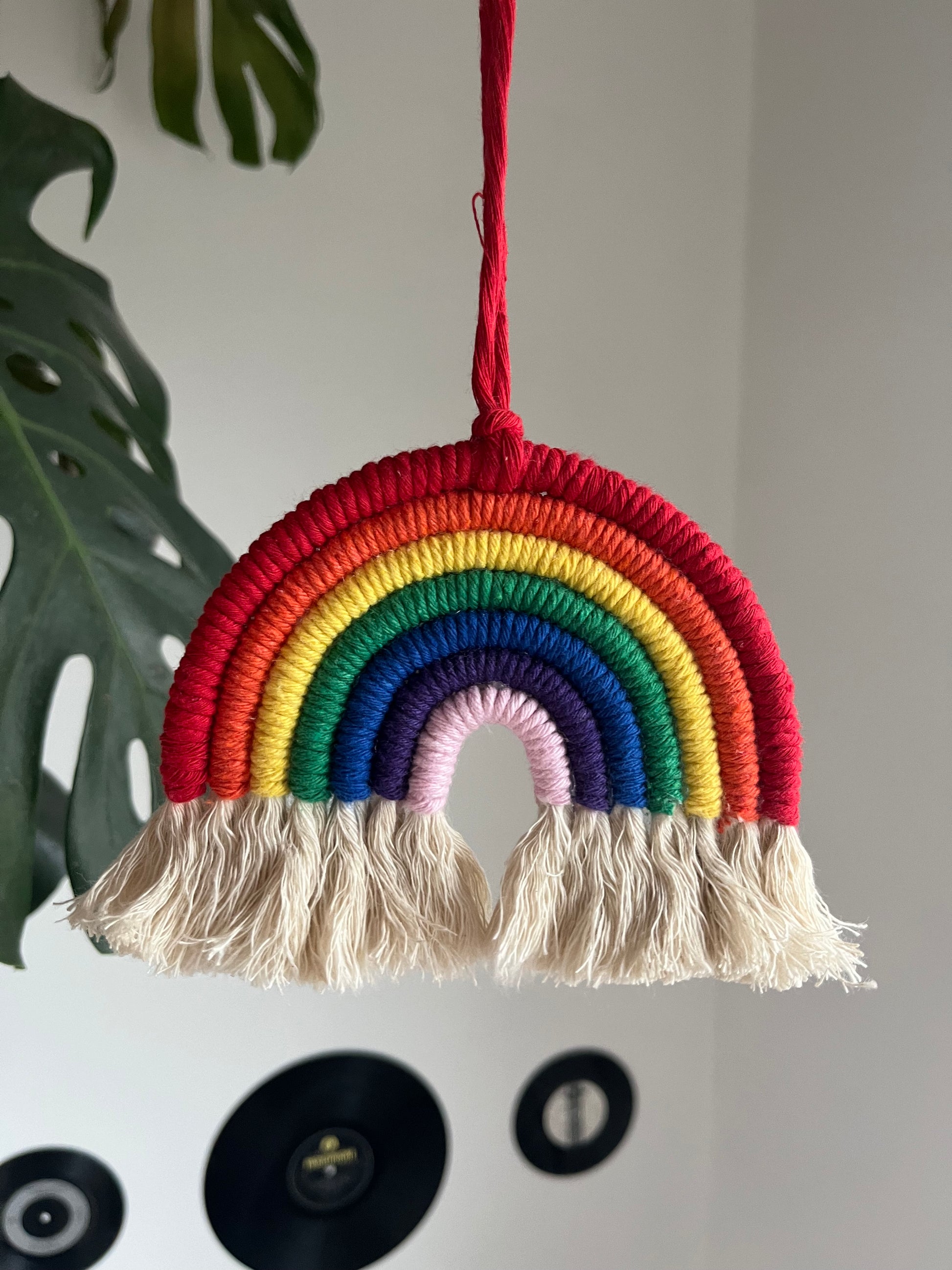 A handmade macrame rainbow wall hanging with vibrant colors, made from recycled cotton yarn, with a red hanging cord and white tassels at the bottom.