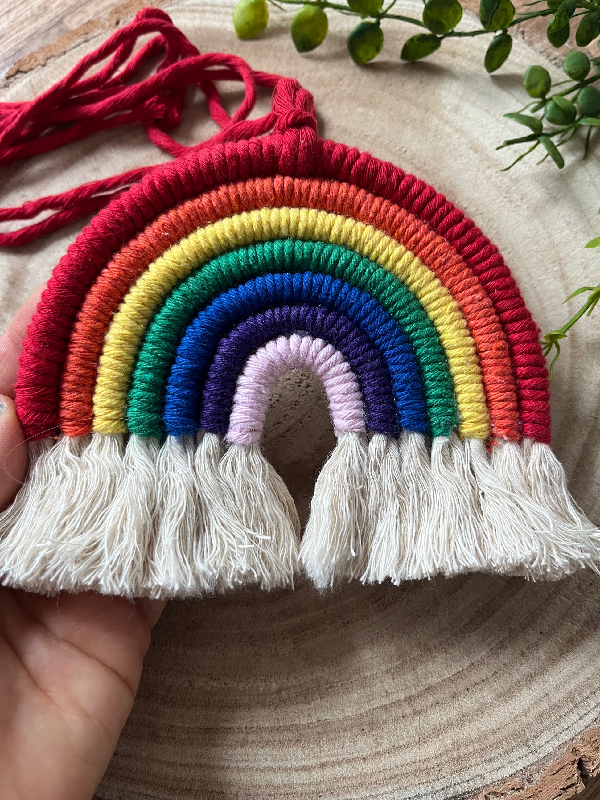A hand holding a handmade macrame rainbow wall hanging with vibrant colours, made from recycled cotton yarn, with a red hanging cord and white tassels at the bottom.