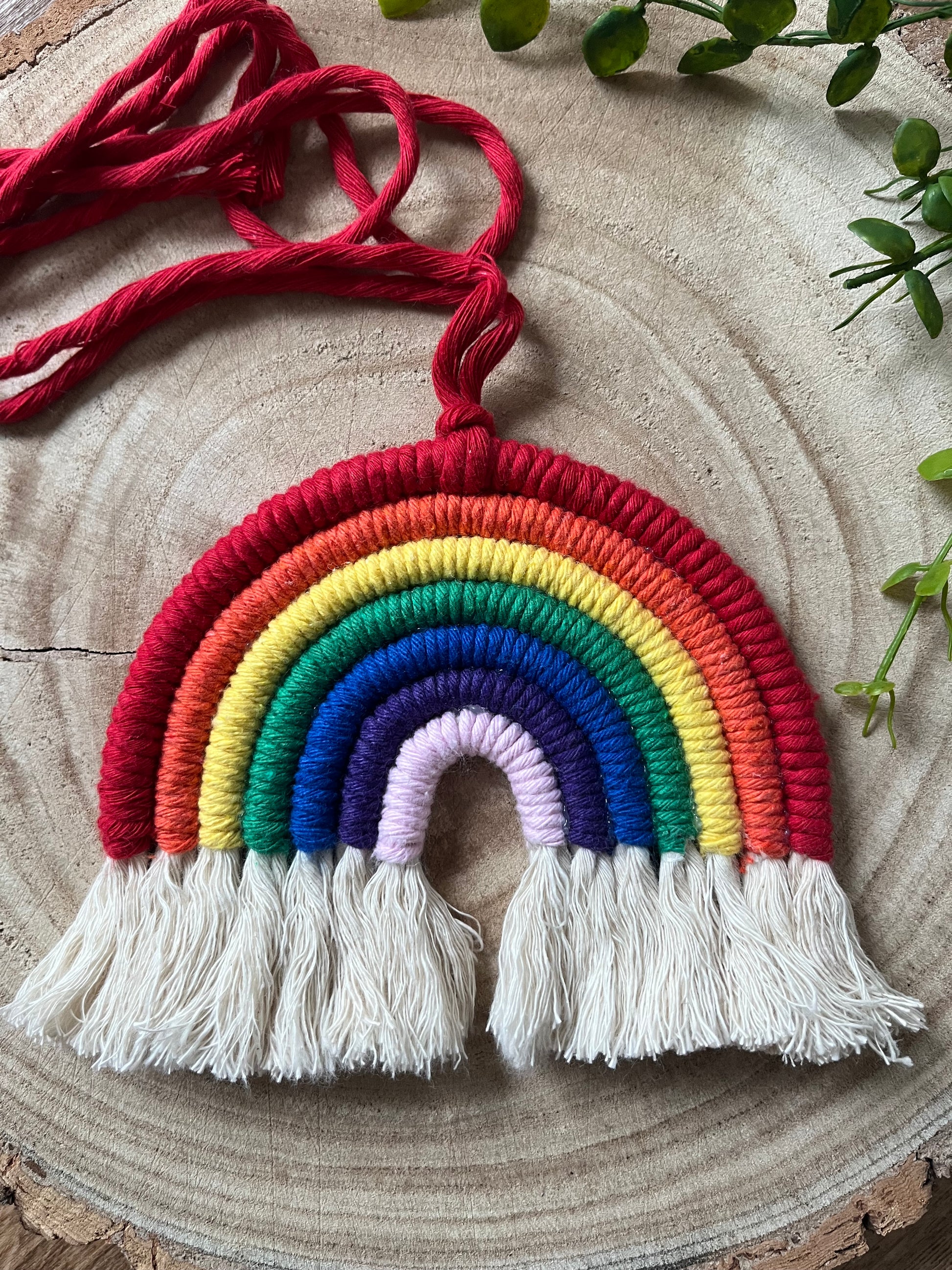 A handmade macrame rainbow wall hanging with vibrant colours, made from recycled cotton yarn, with a red hanging cord and white tassels at the bottom on a wooden board.