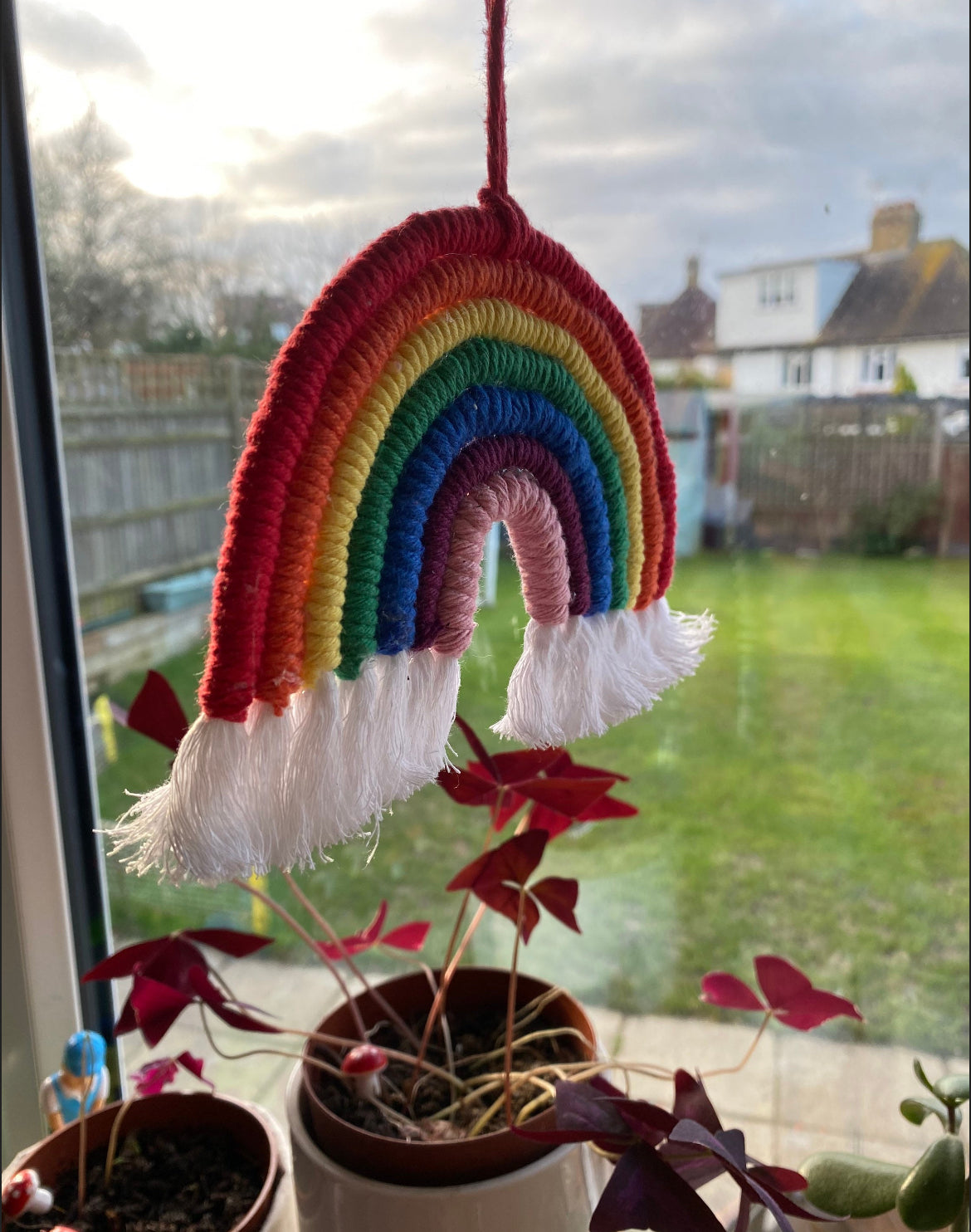 Close up of a handmade macrame rainbow wall hanging with vibrant colours, made from recycled cotton yarn, with a red hanging cord and white tassels at the bottom hanging in a kitchen window.