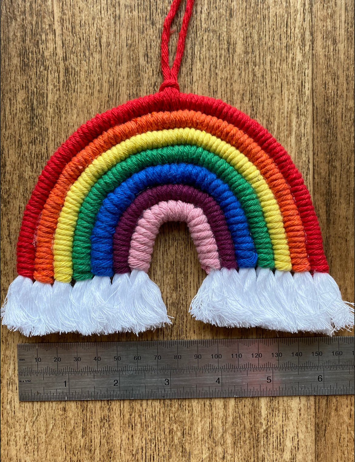 A handmade macrame rainbow wall hanging with vibrant colours, made from recycled cotton yarn, with a red hanging cord and white tassels at the bottom on a wooden floor with a metal ruler underneath it horizontally.