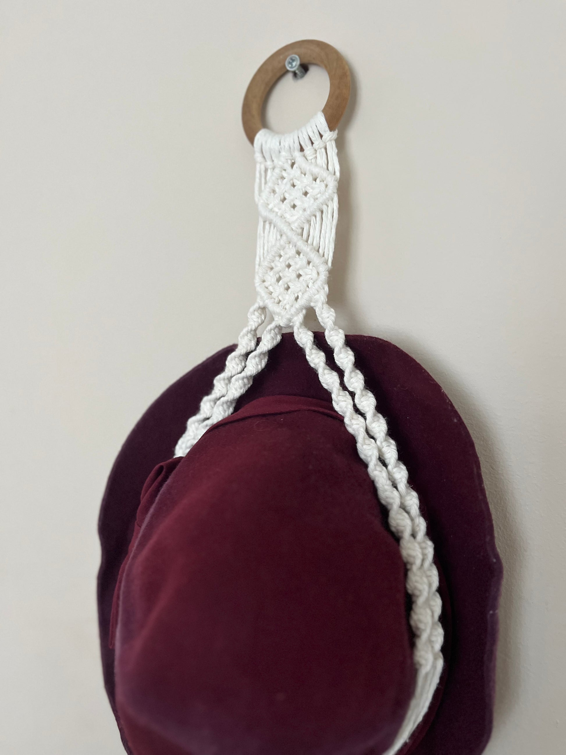 A dark red hat is suspended inside a macrame hat holder, made with natural coloured yarn and a wooden ring, against a white wall.