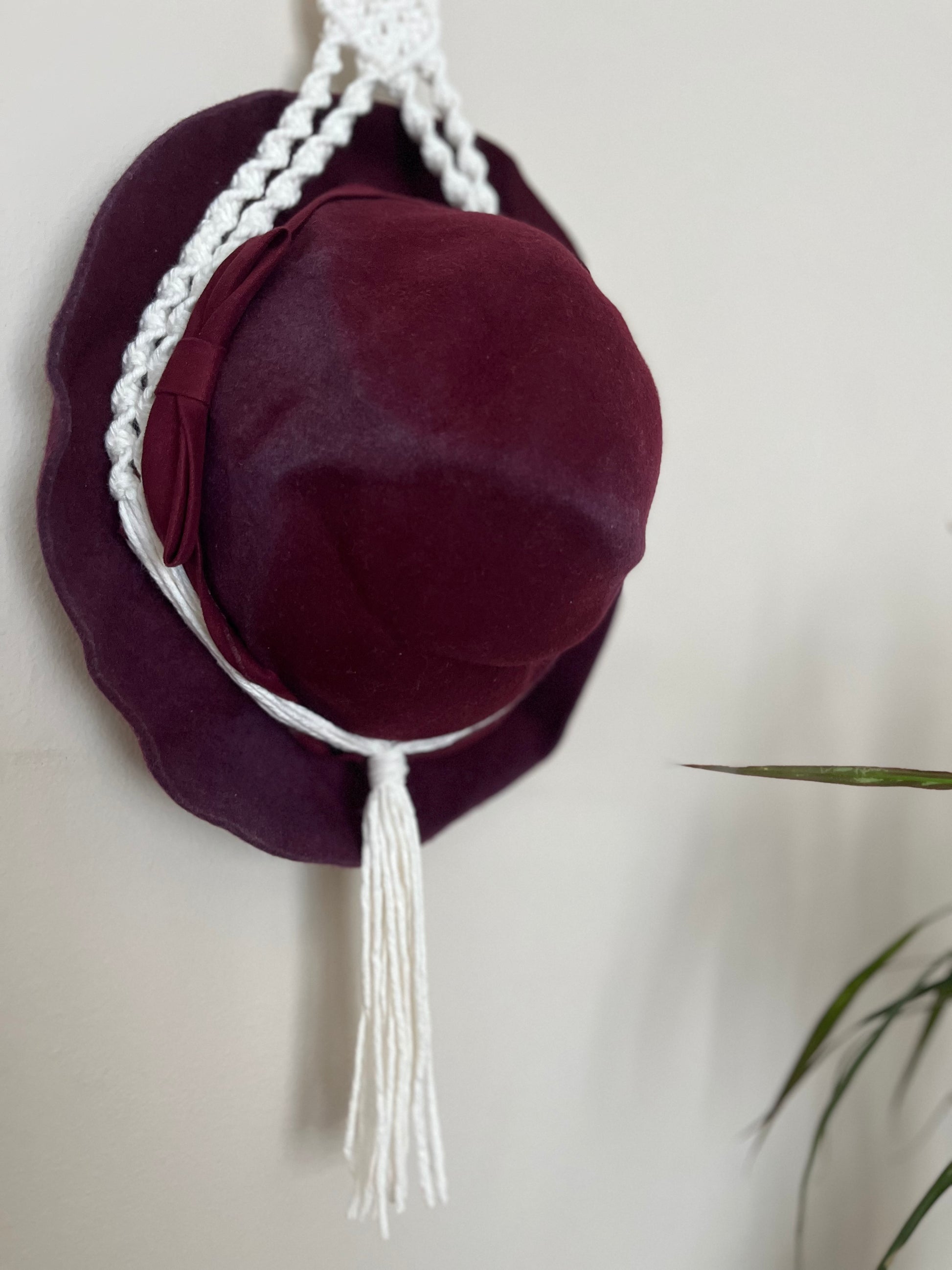 A dark red hat is suspended inside a macrame hat holder, made with natural coloured yarn against a white wall.