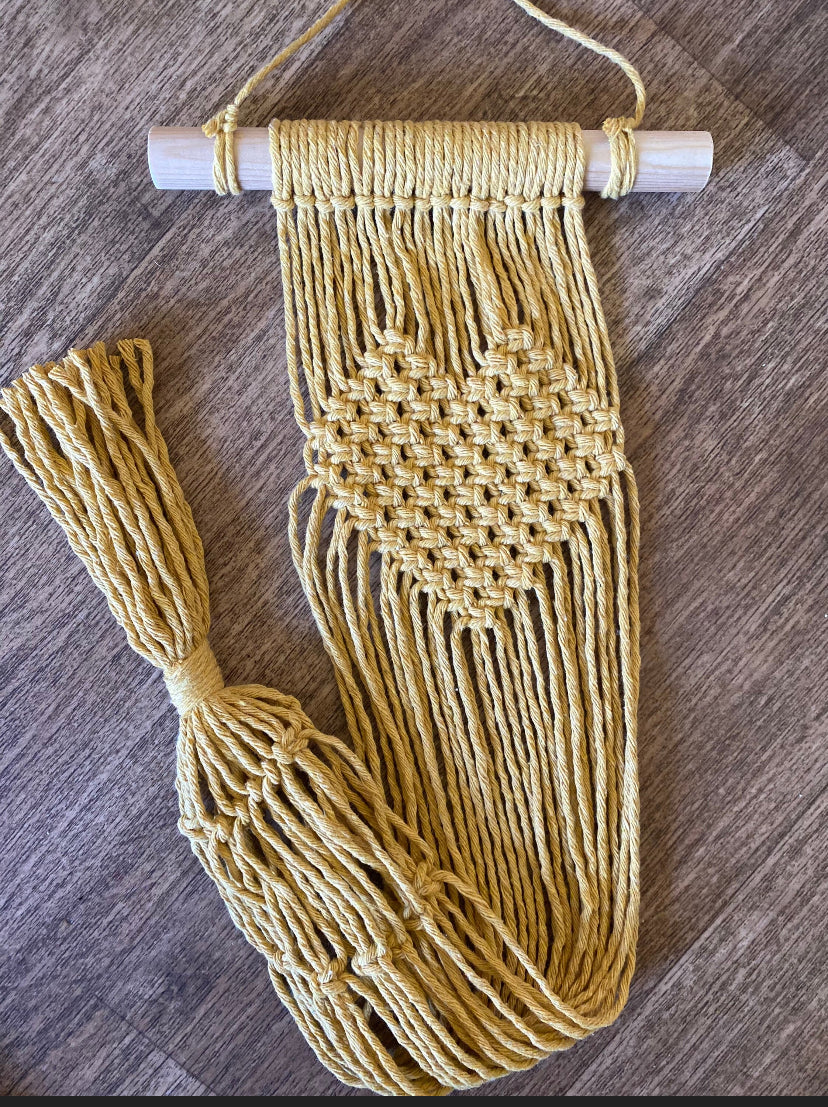 A macrame wall hanging plant holder made with mustard yellow yarn on a wooden dowel with knotted heart detail laid out on a wooden floor.