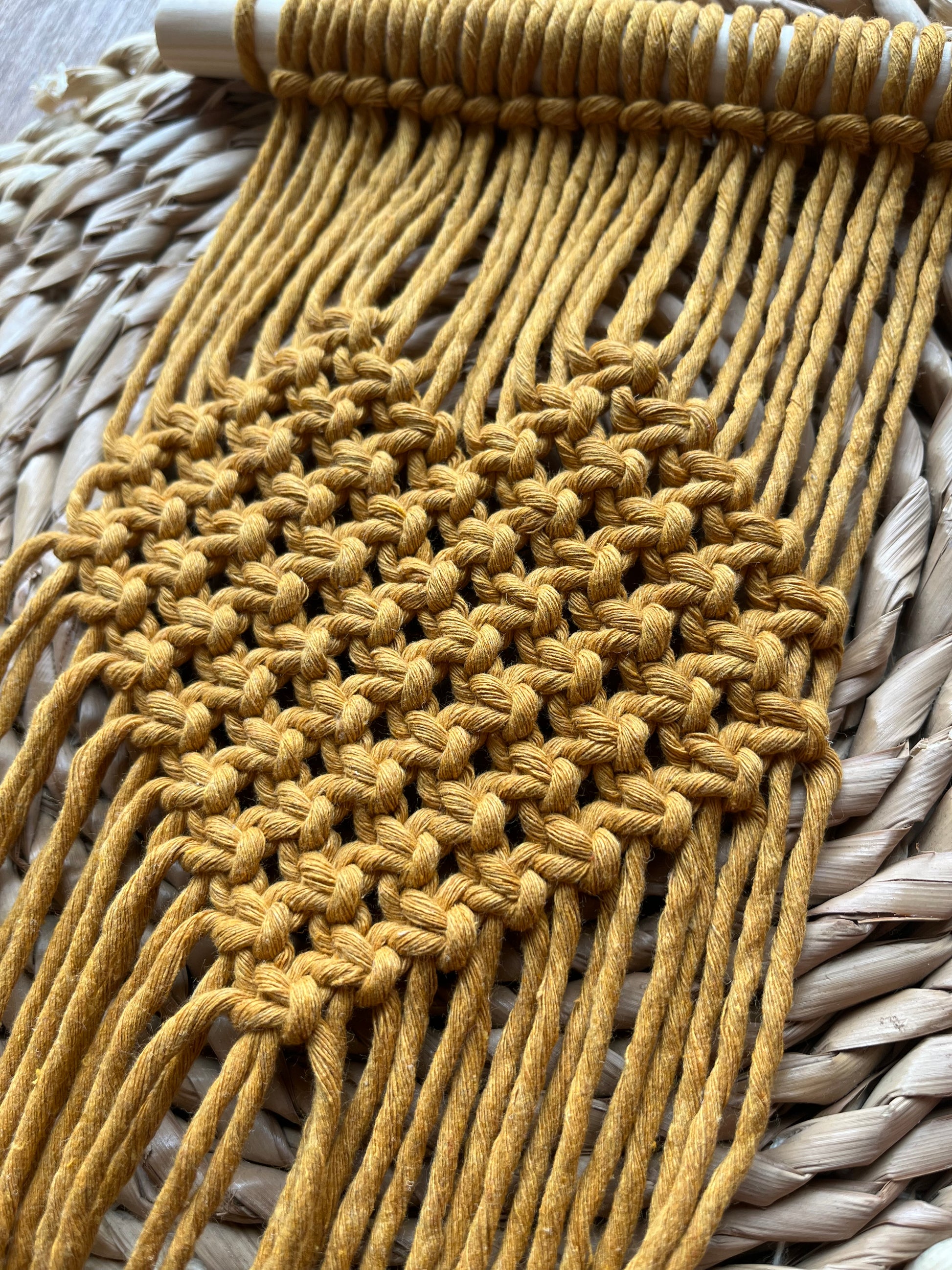 Close up of a mustard yellow macrame wall hanging plant holder with knotted heart detail on a wooden dowel, laid out on a wicker mat.