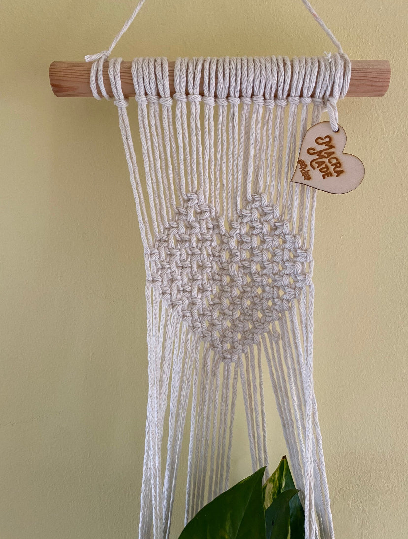 Close up of a natural macrame wall hanging plant holder with knotted heart detail on a wooden dowel against a pale yellow painted wall.
