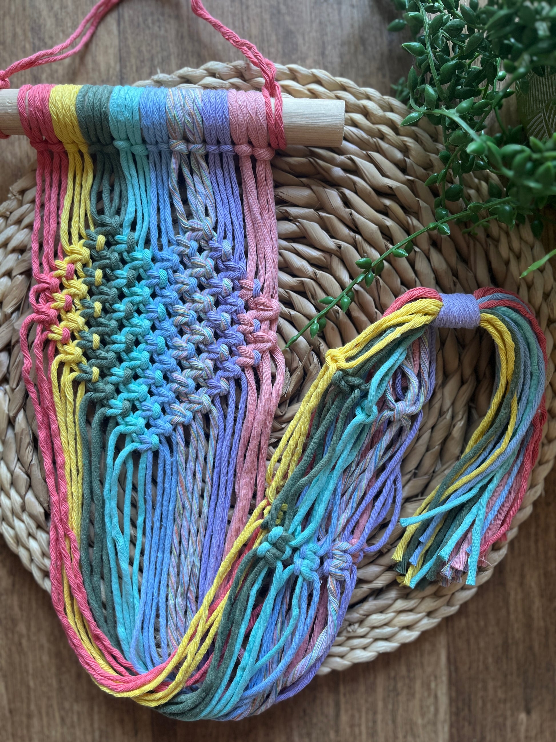 Pastel coloured macrame wall hanging plant holder with knotted heart detail on a wooden dowel, laid out on a wicker mat.