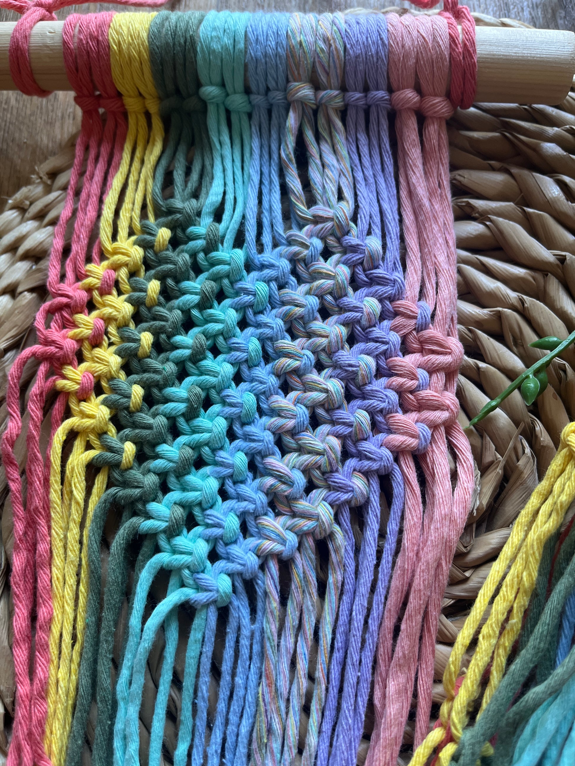 Close up of a pastel coloured macrame wall hanging plant holder with knotted heart detail on a wooden dowel, laid out on a wicker mat.