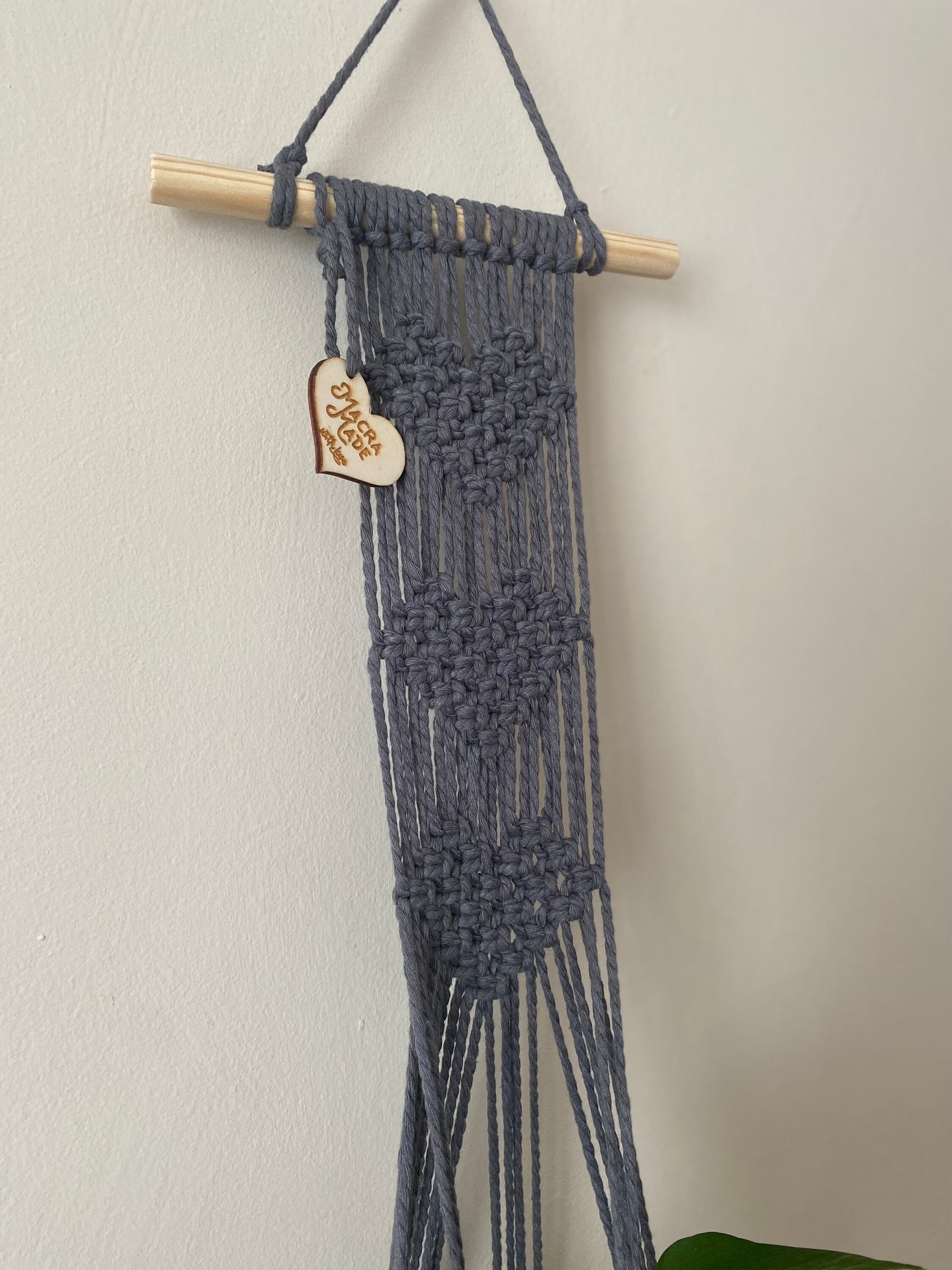 Close up of a macrame wall hanging plant hanger with heart detail, made with slate grey yarn on a small wooden dowel.
