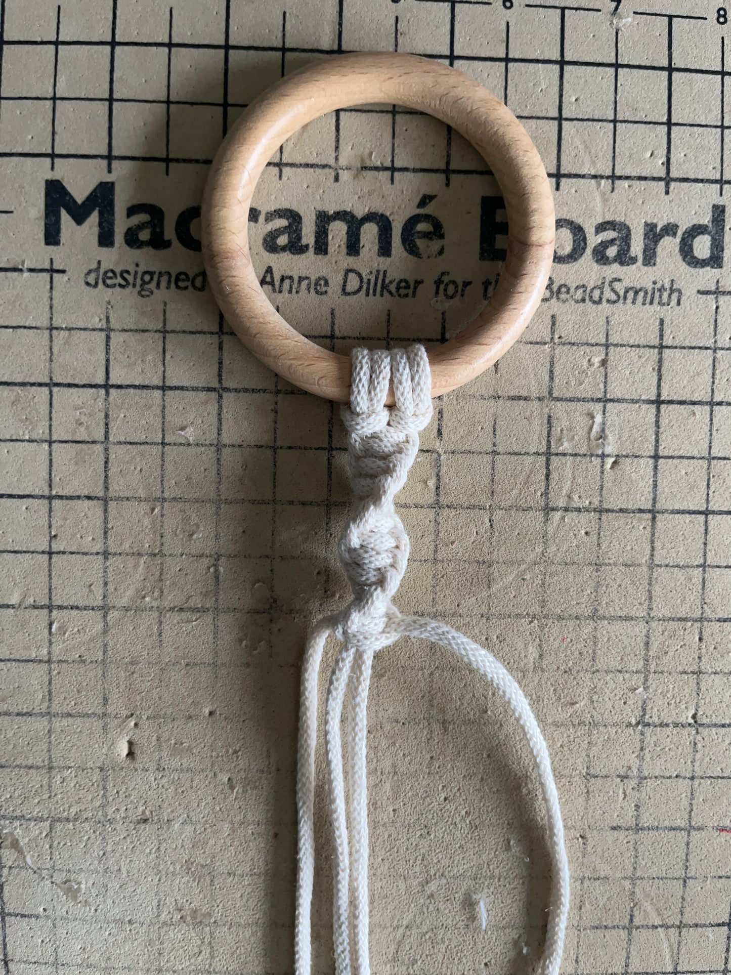 A wooden ring on a checkered macrame board, with natural yarn attached to the ring in a spiral pattern