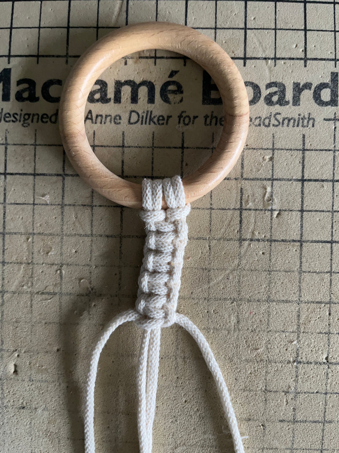 A wooden ring on a checkered macrame board, with natural yarn attached to the ring.