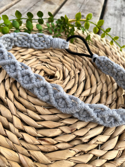 A grey knotted headband made from recycled cotton cord with an elastic finish, displayed on a woven placemat with green plant vines.