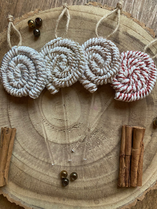 Four handmade macrame christmas decorations in the shape of lollipops, made with metallic yarn and clear acrylic sticks with a jute hanging loop at the top, displayed next to each other on a wooden board, surround by cinnamon sticks and gold beads for decoration.