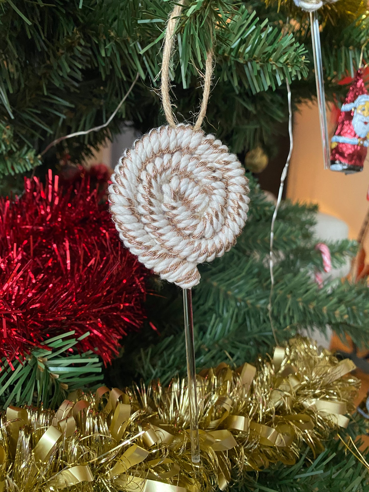 A handmade macrame christmas decoration in the shape of a lollipop, made with metallic yarn and clear acrylic stick with a jute hanging loop at the top, displayed hanging amongst tinsel and greenery on an artificial christmas tree.