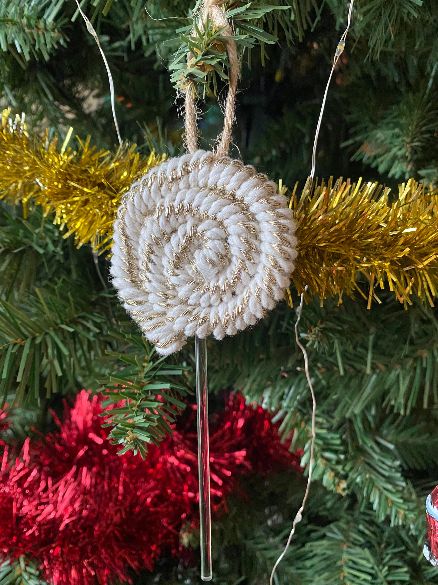 A handmade macrame christmas decoration in the shape of a lollipop, made with gold and white metallic yarn and clear acrylic stick with a jute hanging loop at the top, displayed hanging amongst tinsel and greenery on an artificial christmas tree.