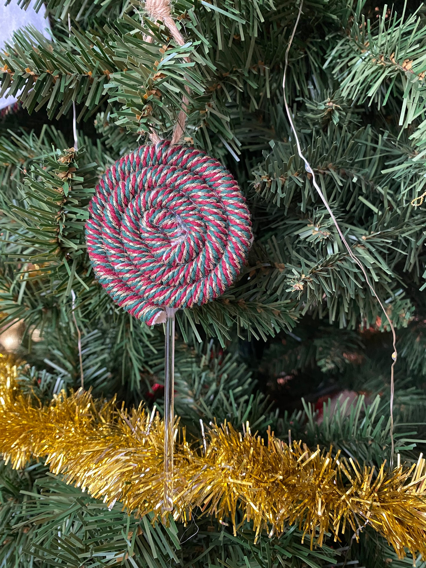 A handmade macrame christmas decoration in the shape of a lollipop, made with red and green metallic yarn and clear acrylic stick with a jute hanging loop at the top, displayed hanging amongst gold tinsel and greenery on an artificial christmas tree.