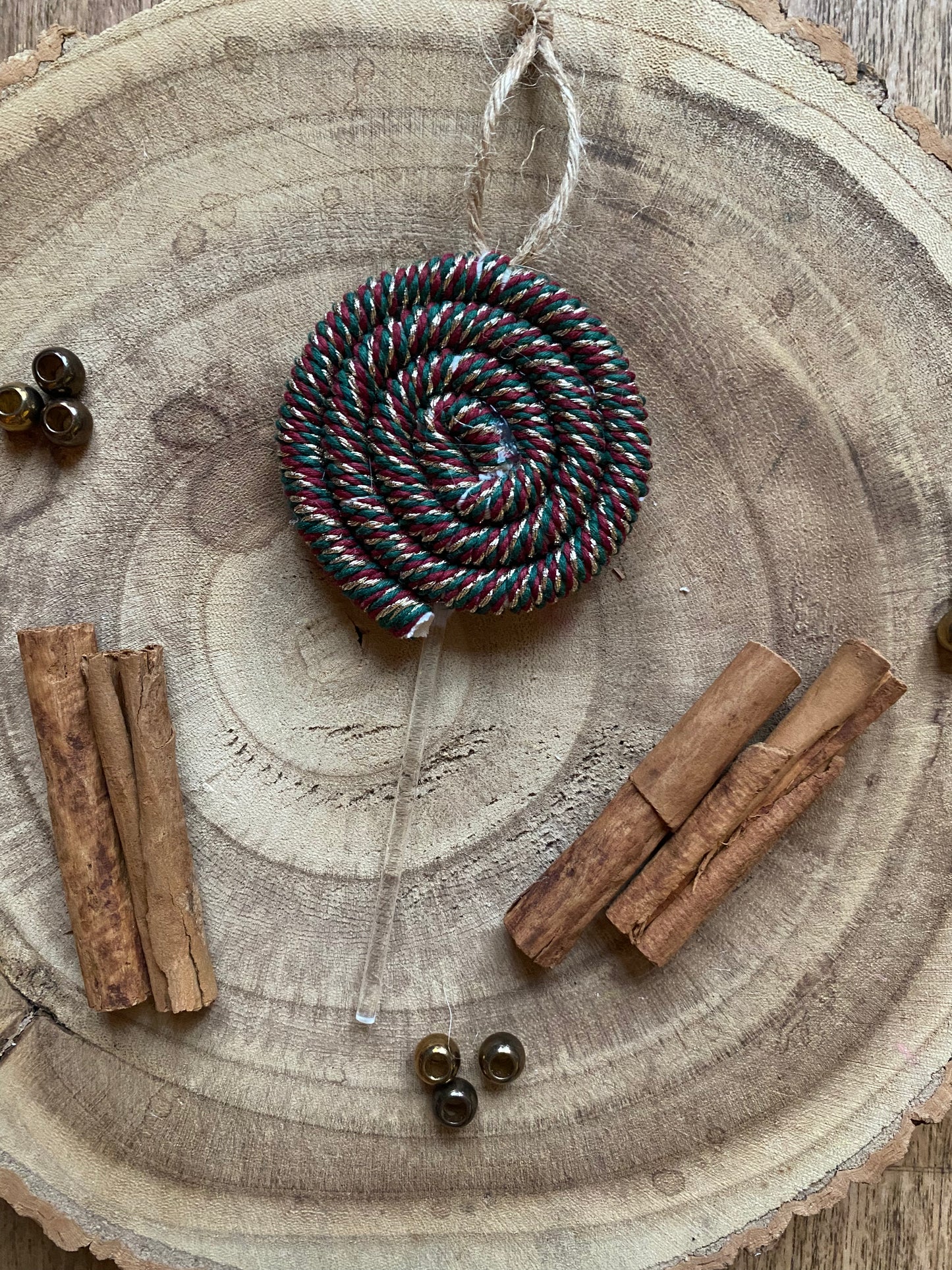 A handmade macrame christmas decoration in the shape of a lollipop, made with red green and gold metallic yarn and clear acrylic stick with a jute hanging loop at the top, displayed on a wooden board with cinnamon sticks and gold beads for decoration.
