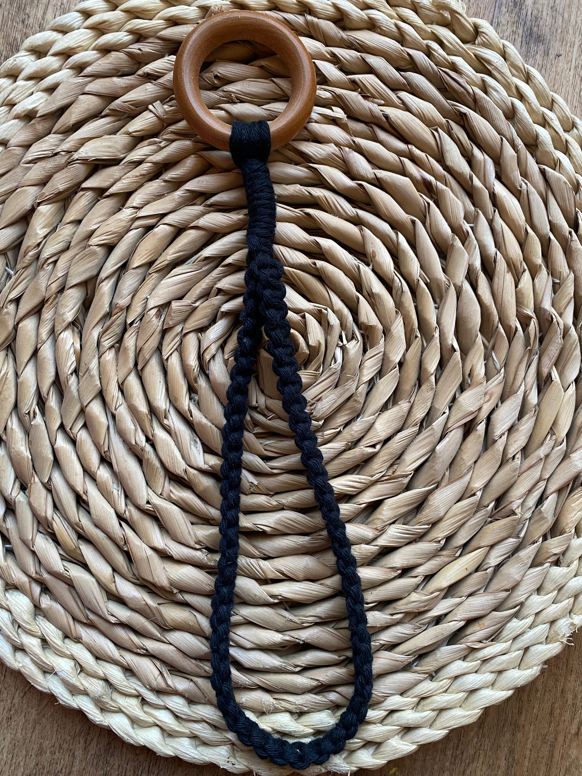 A handmade macrame ukulele hanger loop made with black yarn and a wooden ring, laid out across a round wicker mat.