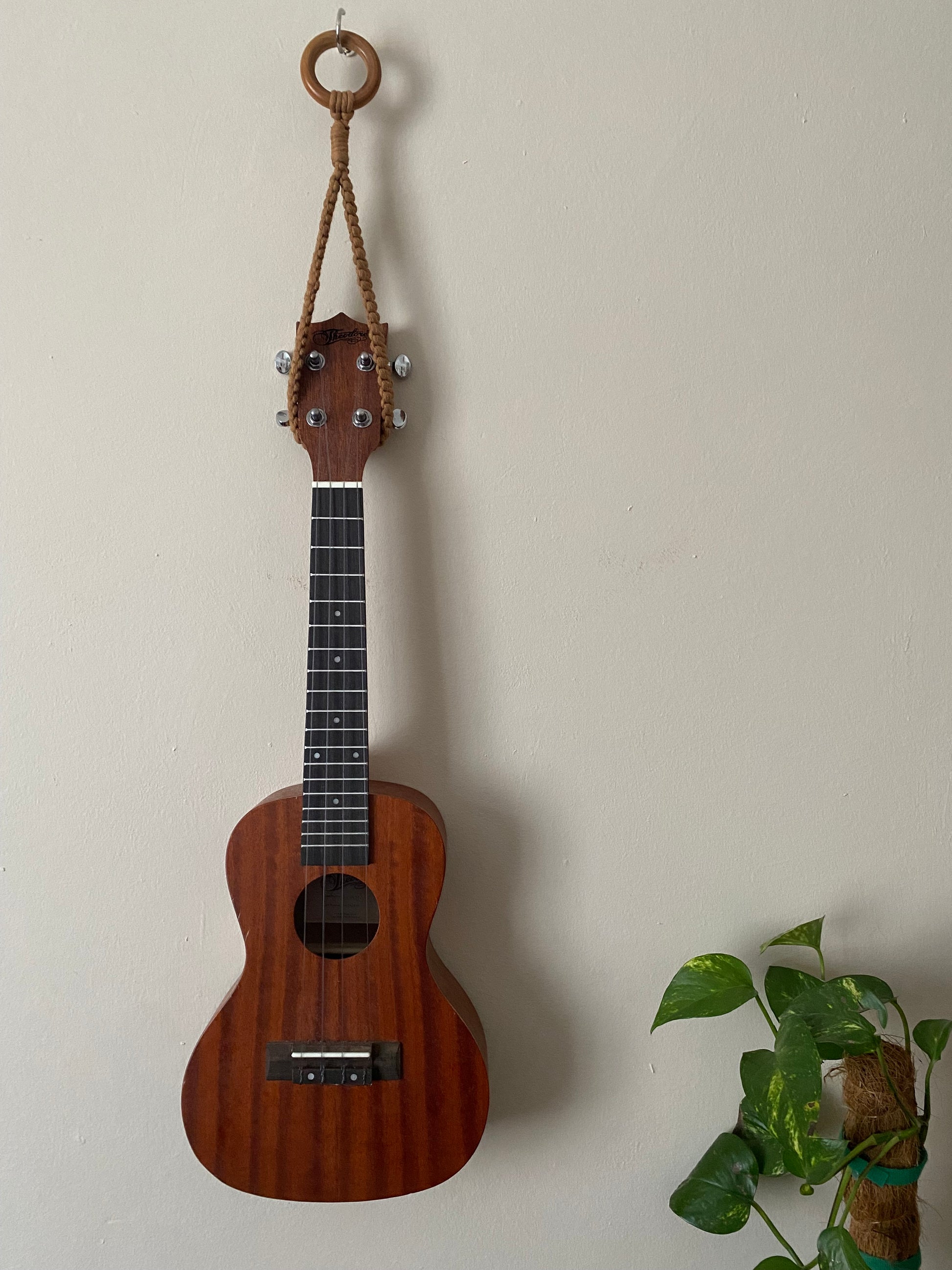 A dark brown ukulele, hanging against a beige wall from a simple macrame knotted loop hanger, hooked around the headstock of the instrument, made with cotton yarn and a wooden ring.